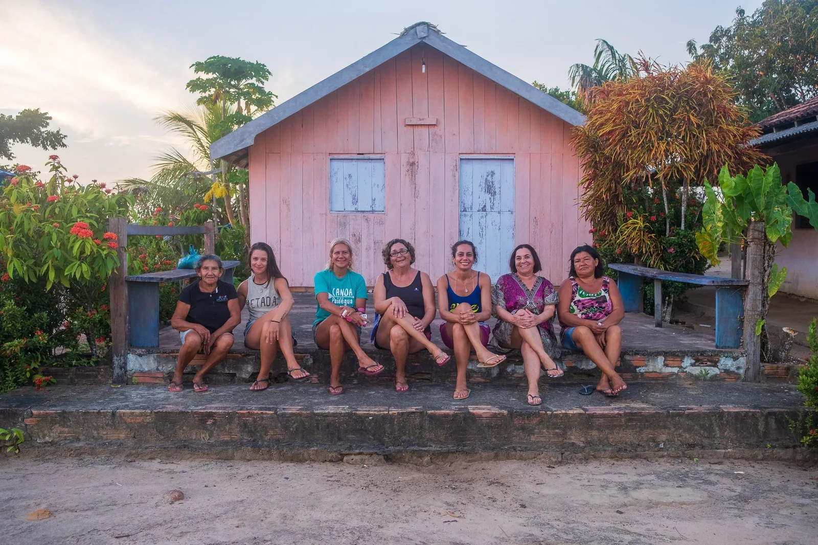 Seven women sitting in a row on a low wall in front of a pink wooden house, flowering plants and trees around it