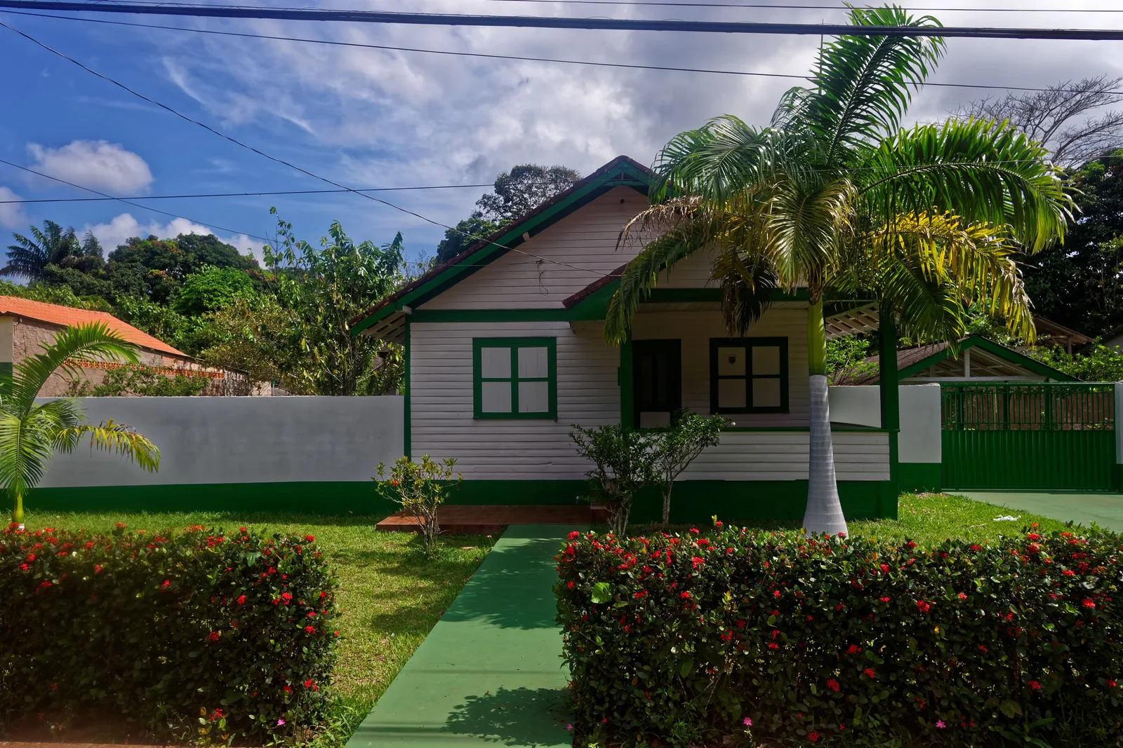 A white wooden bungalow with green trim and a pitched roof behind a clipped hedge and a short green path, a palm tree beside it