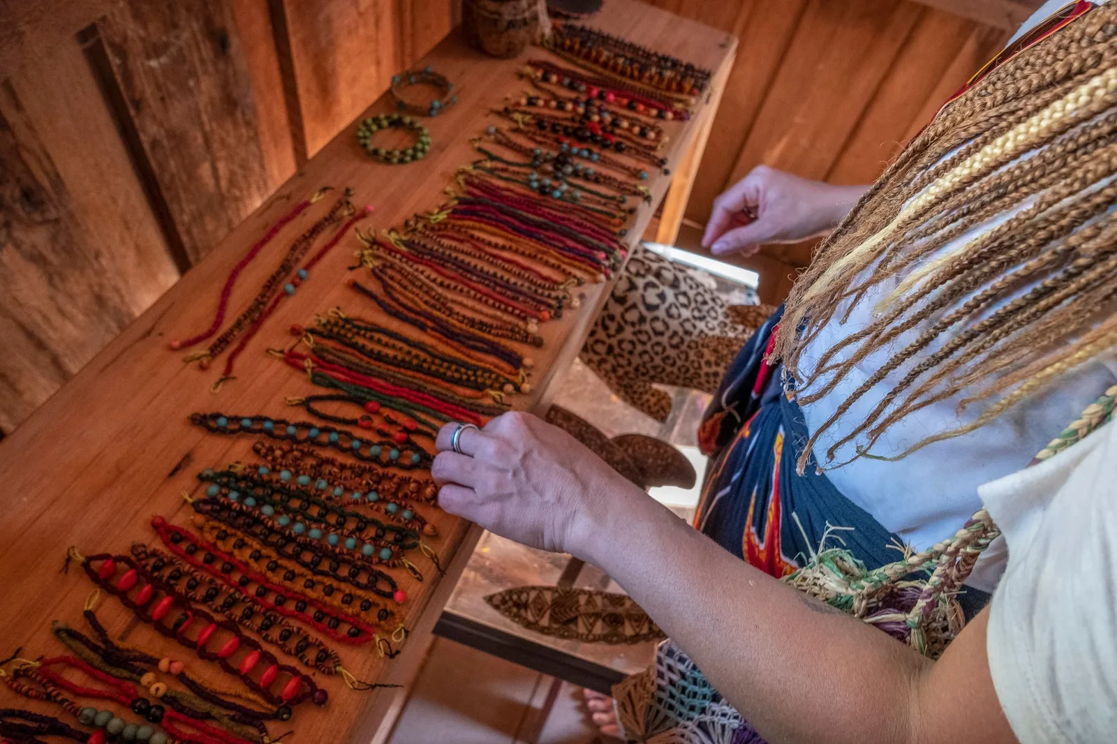 Hands reaching over a wooden counter lined with rows of handmade bracelets and necklaces of red and brown seeds, a woven bag hanging beside them