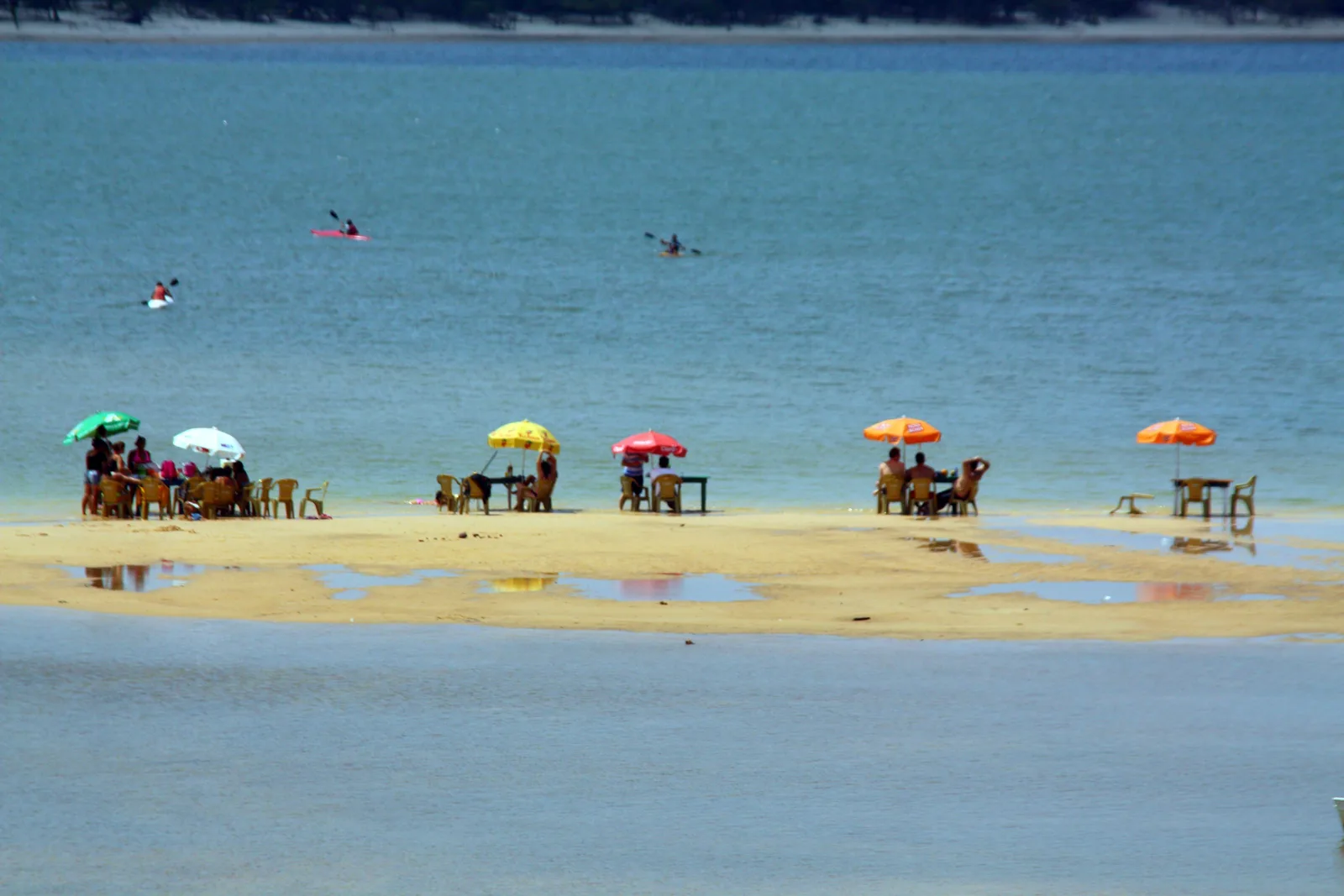 Tables and bright umbrellas set out on a strip of sand in the middle of clear turquoise river water, a few kayaks on the water beyond