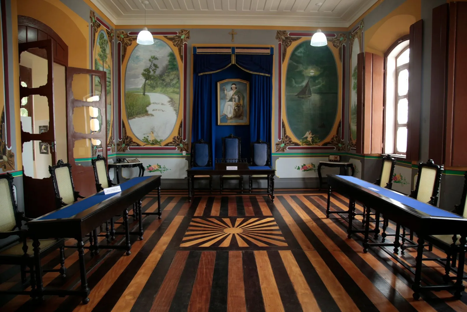 A formal room with a striped inlaid wooden floor, long blue-topped tables with cane chairs, painted landscape panels on the walls and a portrait under a blue canopy at the far end