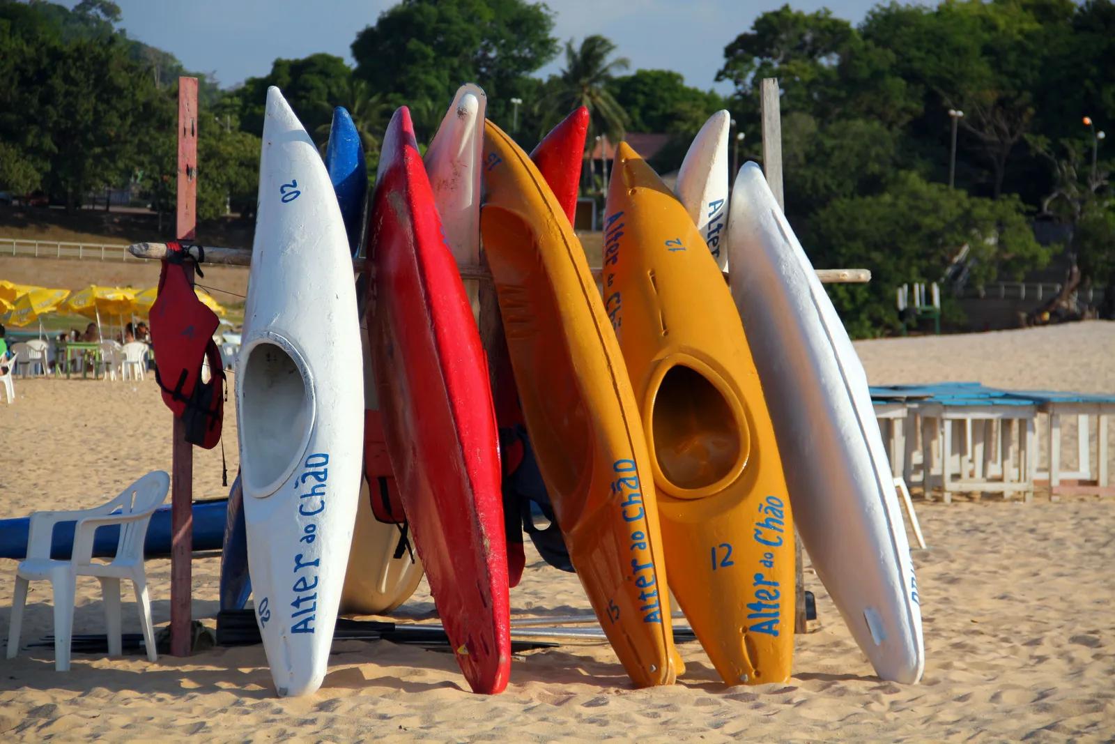 A row of white, red and yellow kayaks with “Alter do Chão” painted on them, standing on end against a rack on a sandy beach
