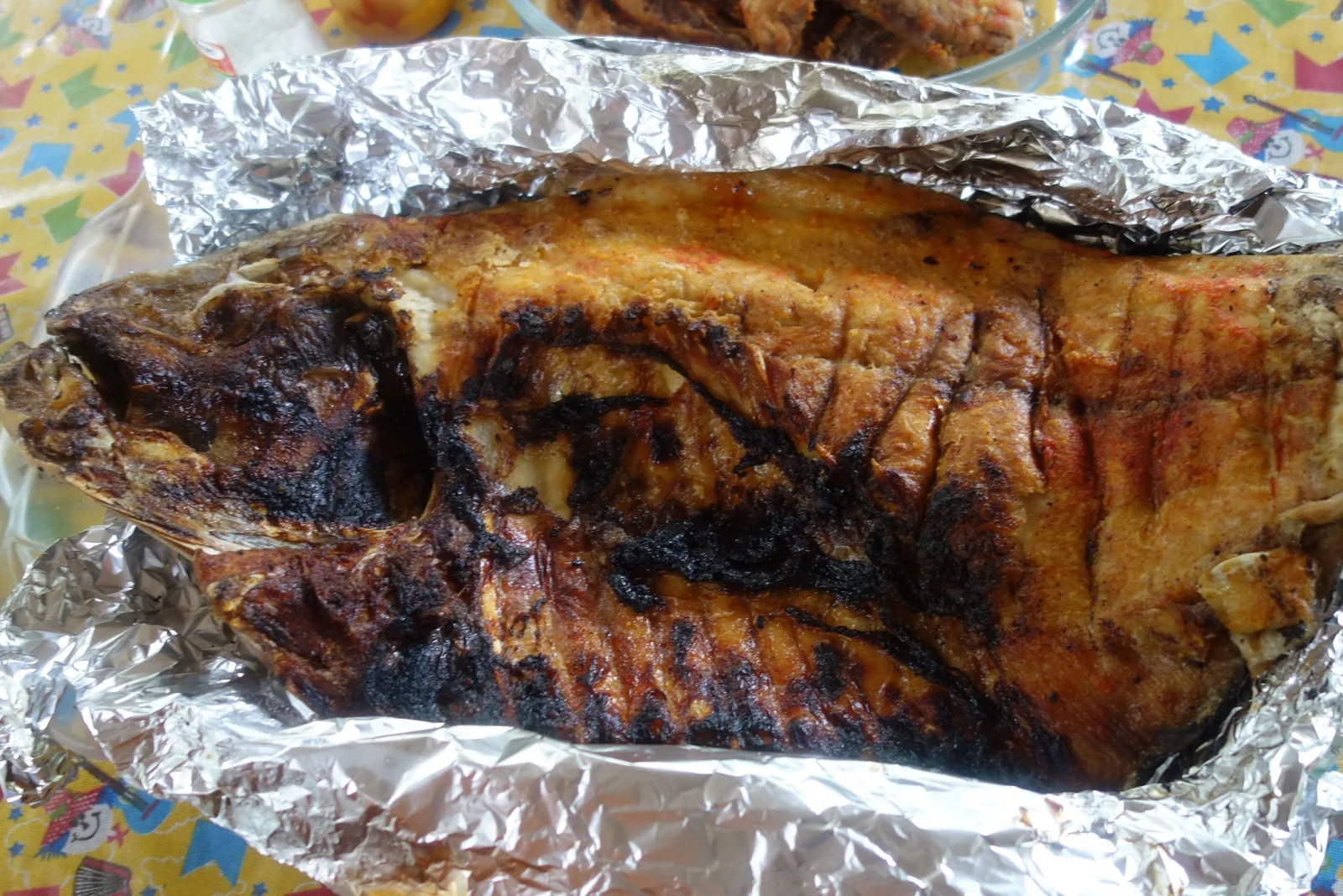 A whole grilled river fish, charred and split open, lying on a sheet of crumpled foil