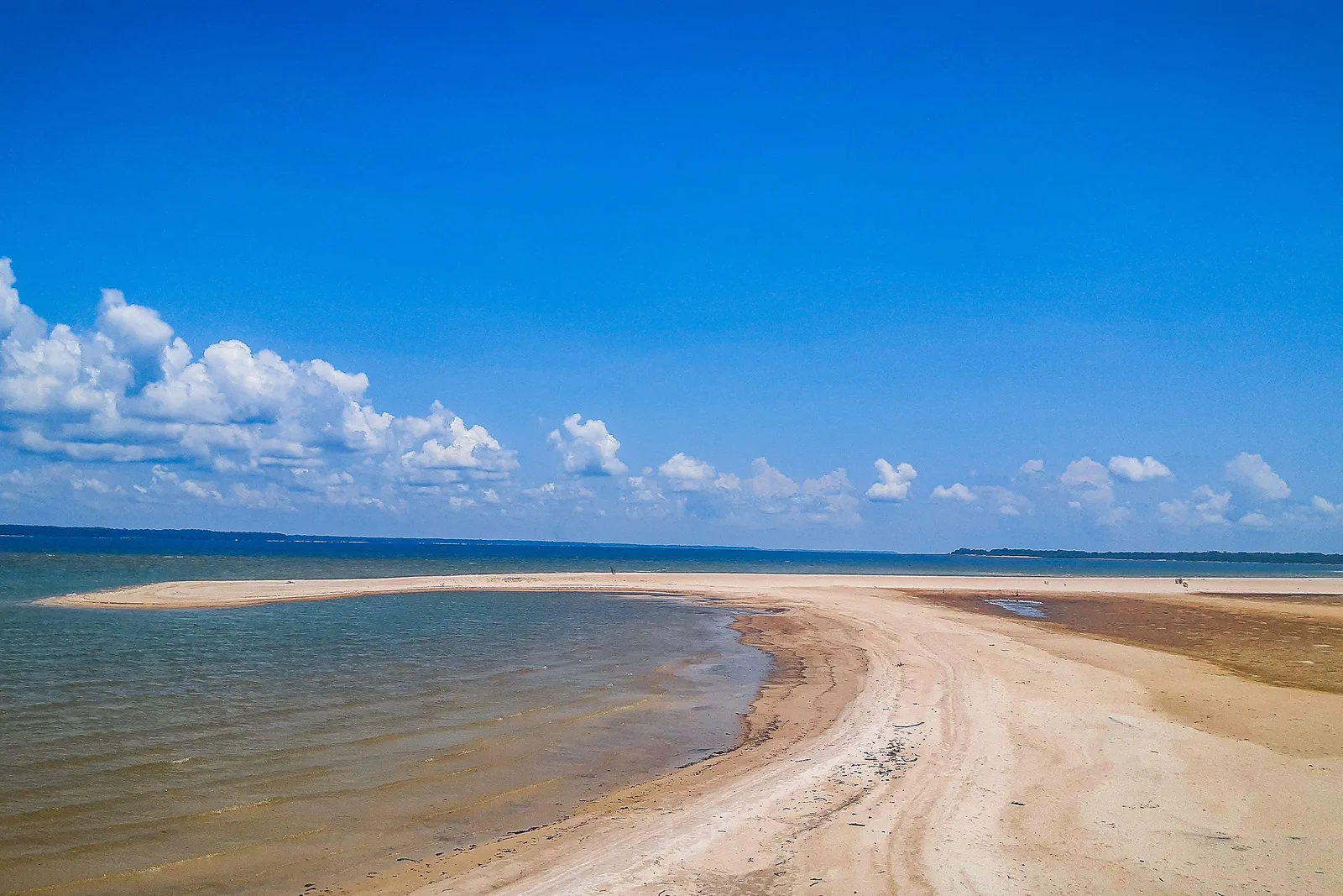 A narrow white sand point curving out into deep blue water under a bright sky with small clouds