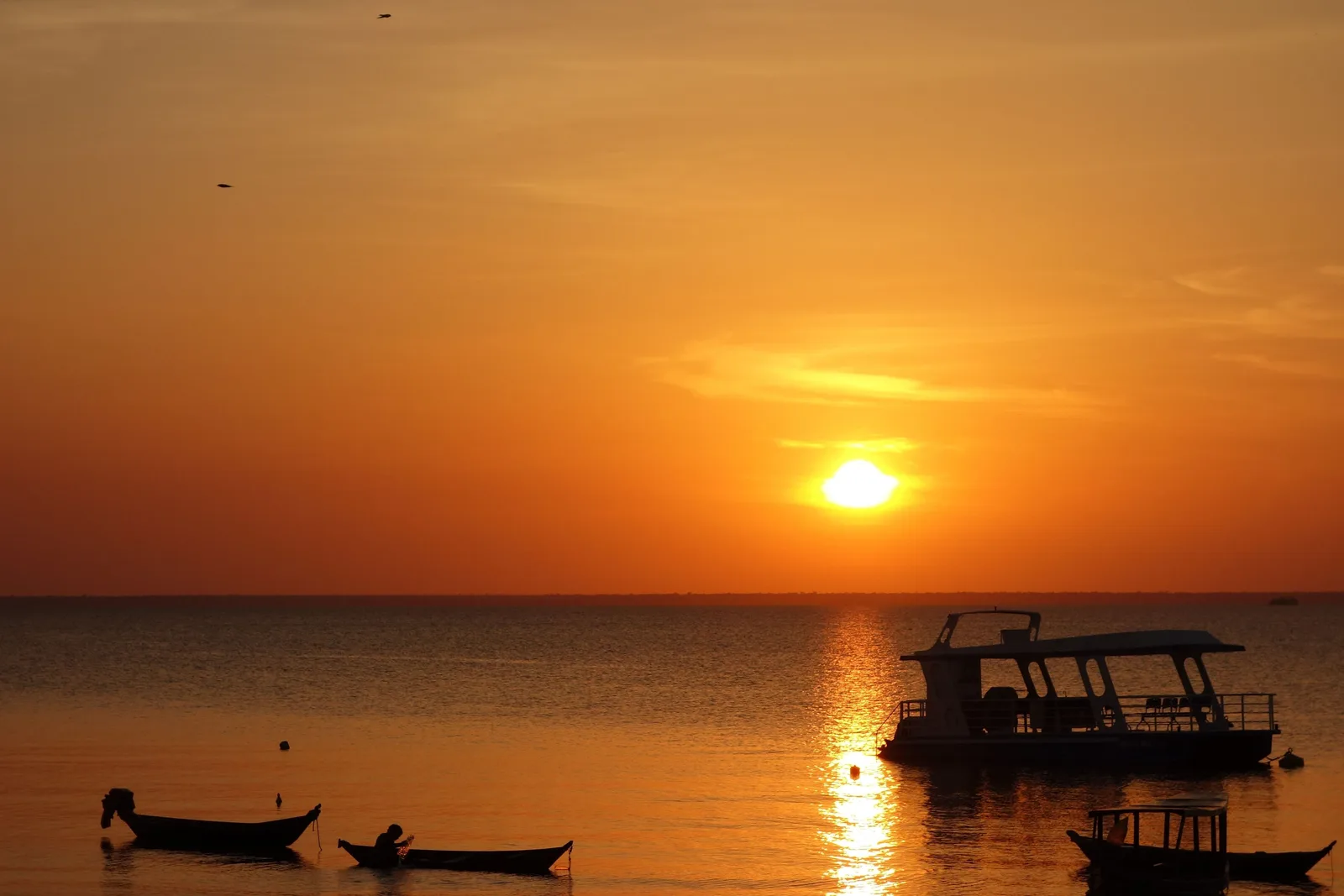 The sun setting in an orange sky over a wide, calm river, a small double-decker riverboat and two canoes silhouetted on the water