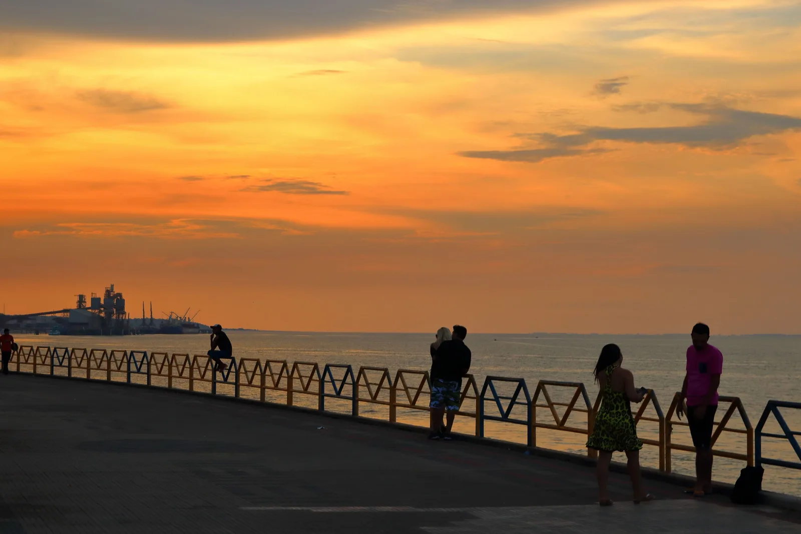 People standing at a railing along a riverside promenade at sunset, silhouetted against an orange and gold sky with a ship on the river