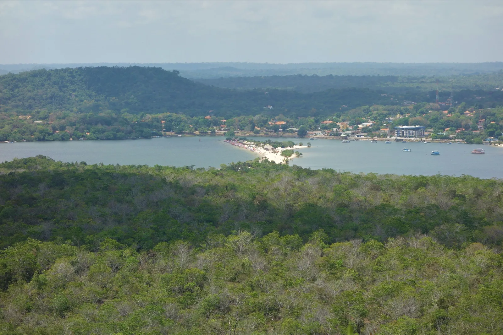 Looking down from a hilltop over dense green forest to a wide bay of blue water, a strip of sand and a village spread along the far shore under flat-topped hills