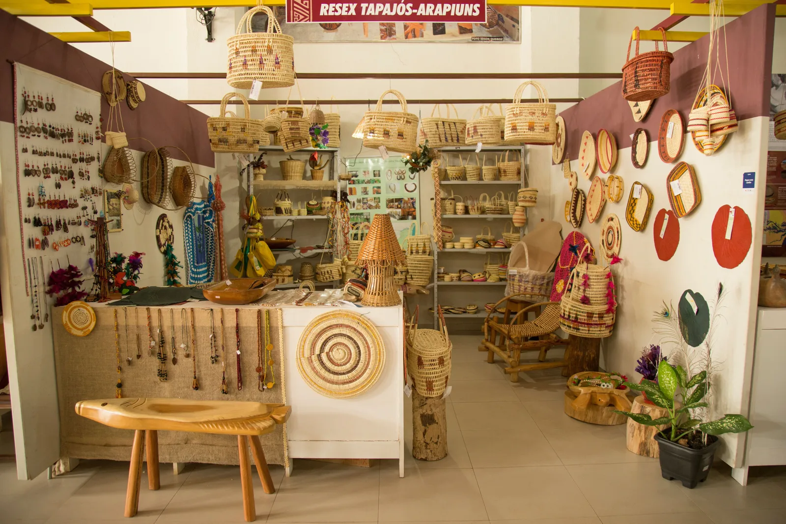 A small craft shop crowded with woven straw baskets, bags and mats, round woven trivets on the walls and a wooden stool in front, under a sign reading “Resex Tapajós-Arapiuns”