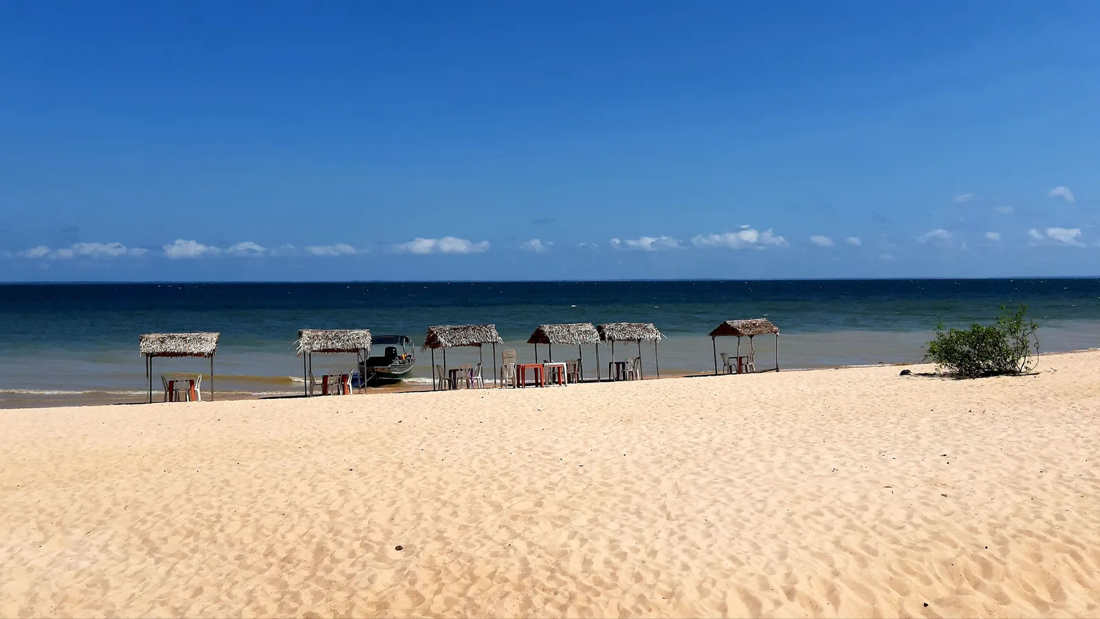 A wide river beach of pale, ridged sand under a deep blue sky, a row of small thatched shelters along the waterline and the river stretching away like a sea