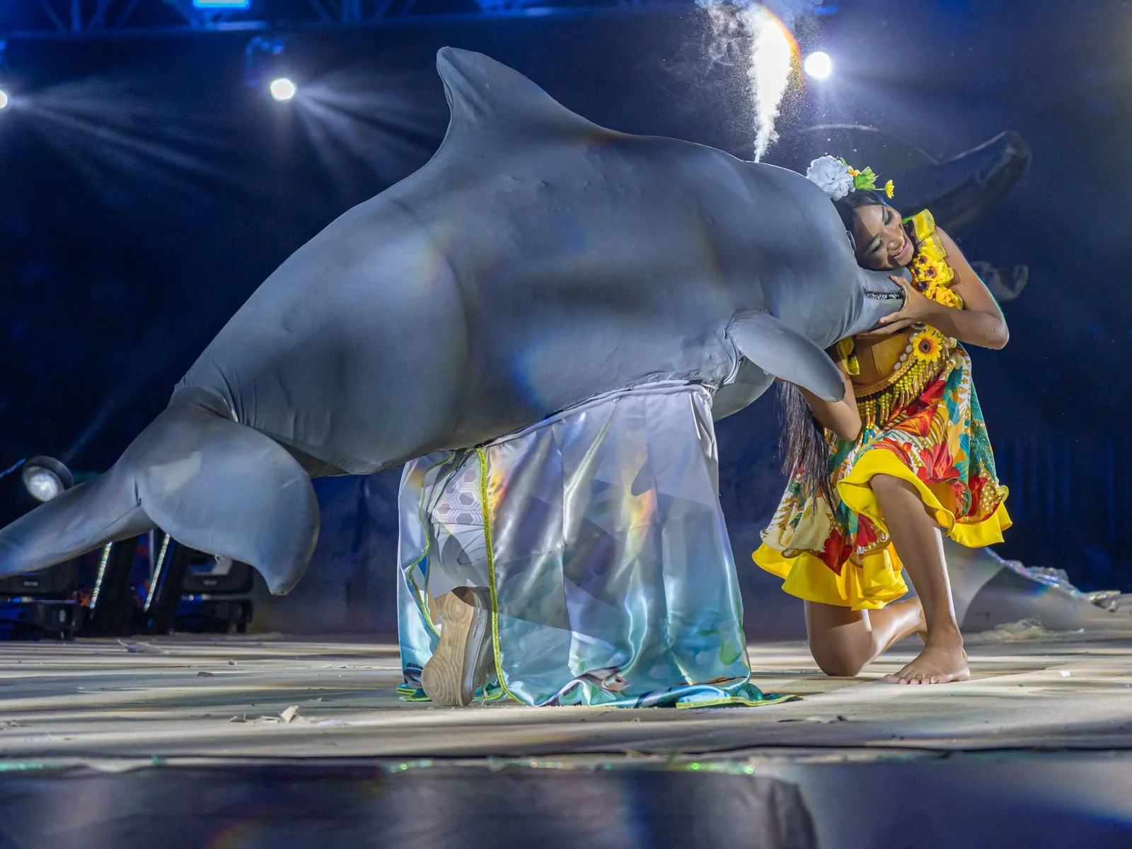 A dancer in a bright yellow dress kneeling on a stage beside a huge gray river-dolphin figure carried by a performer in silver, spotlights and sparks behind