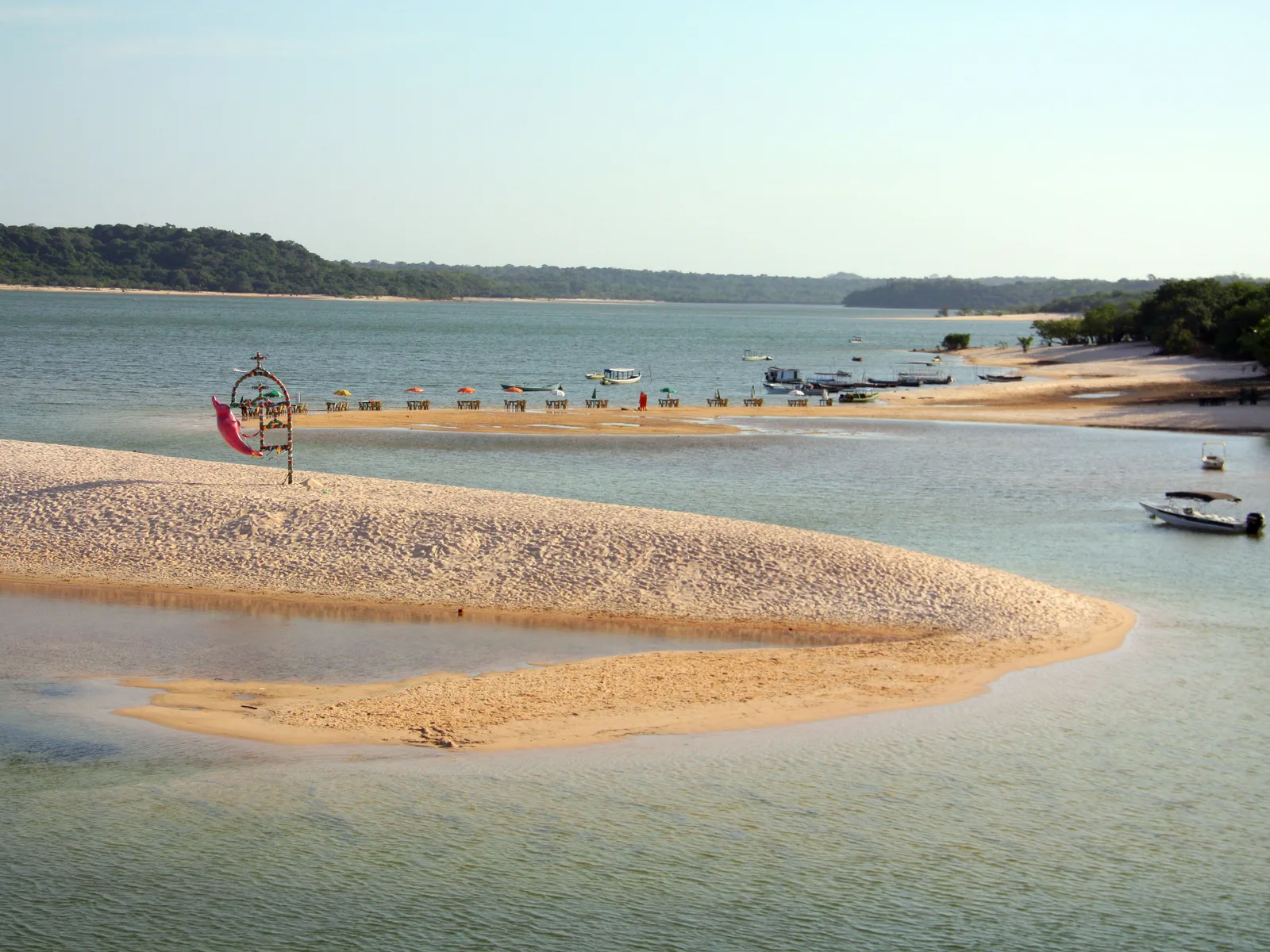 A long tongue of pale sand exposed in shallow, clear river water, a decorated pole on the sand and boats moored off a beach lined with umbrellas behind