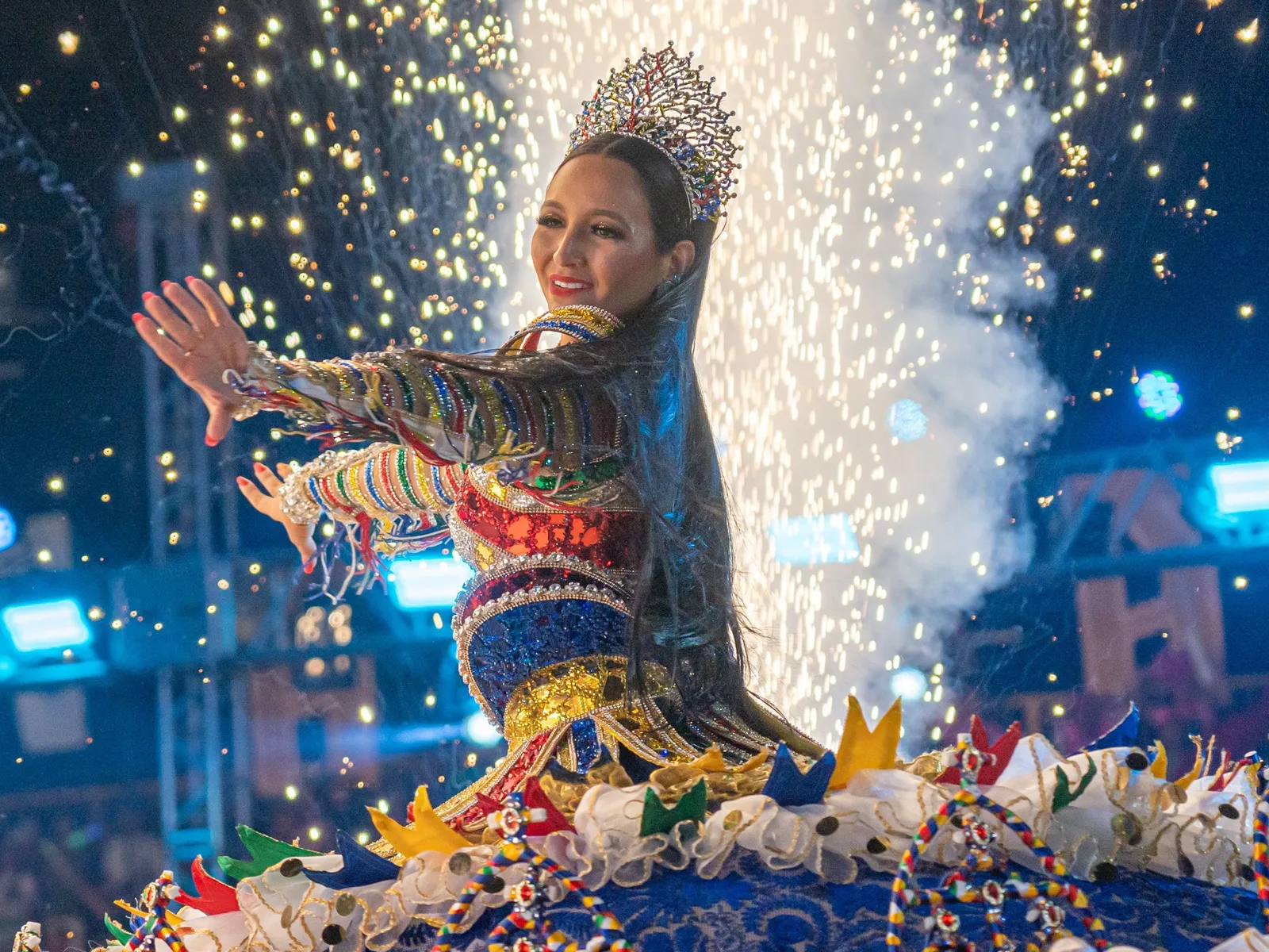 A performer in a jeweled crown and an elaborately beaded costume kneeling with her arms outstretched on a float, sparks and smoke rising behind her on a lit stage at night