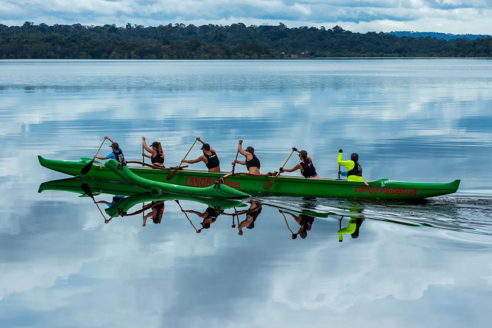 A long green outrigger canoe with six paddlers racing across flat water that mirrors the clouds and the forest on the far bank