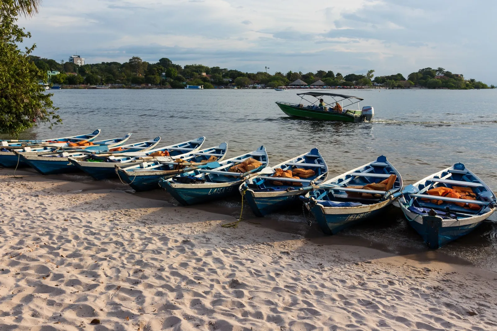 A row of small blue wooden boats pulled up on a sandy beach, a motorboat crossing the river behind them and roofs and a taller building among the trees on the far shore
