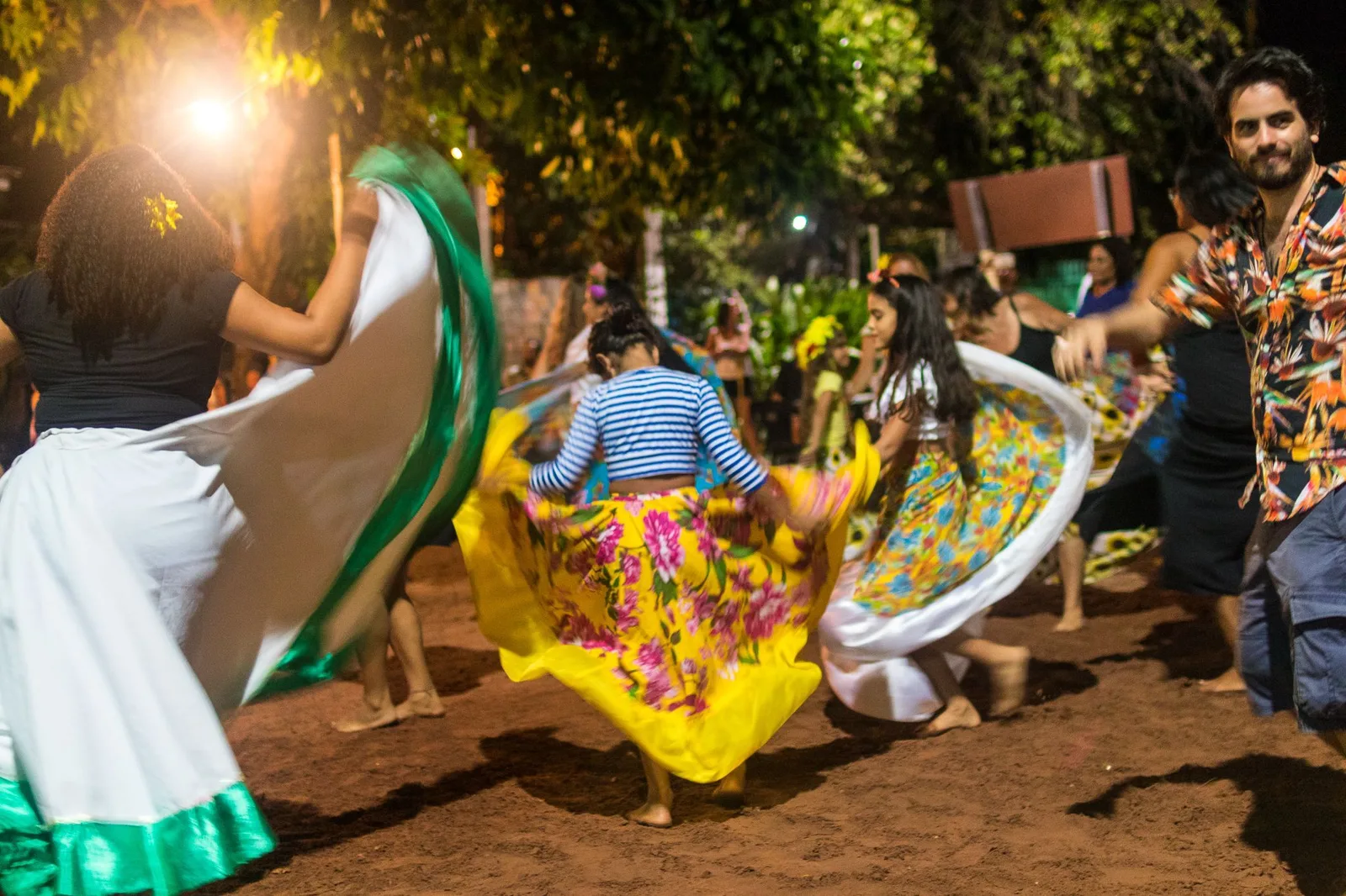 Dancers in long, full yellow and white skirts spinning on a sandy floor at night under string lights, a man in a patterned shirt dancing beside them