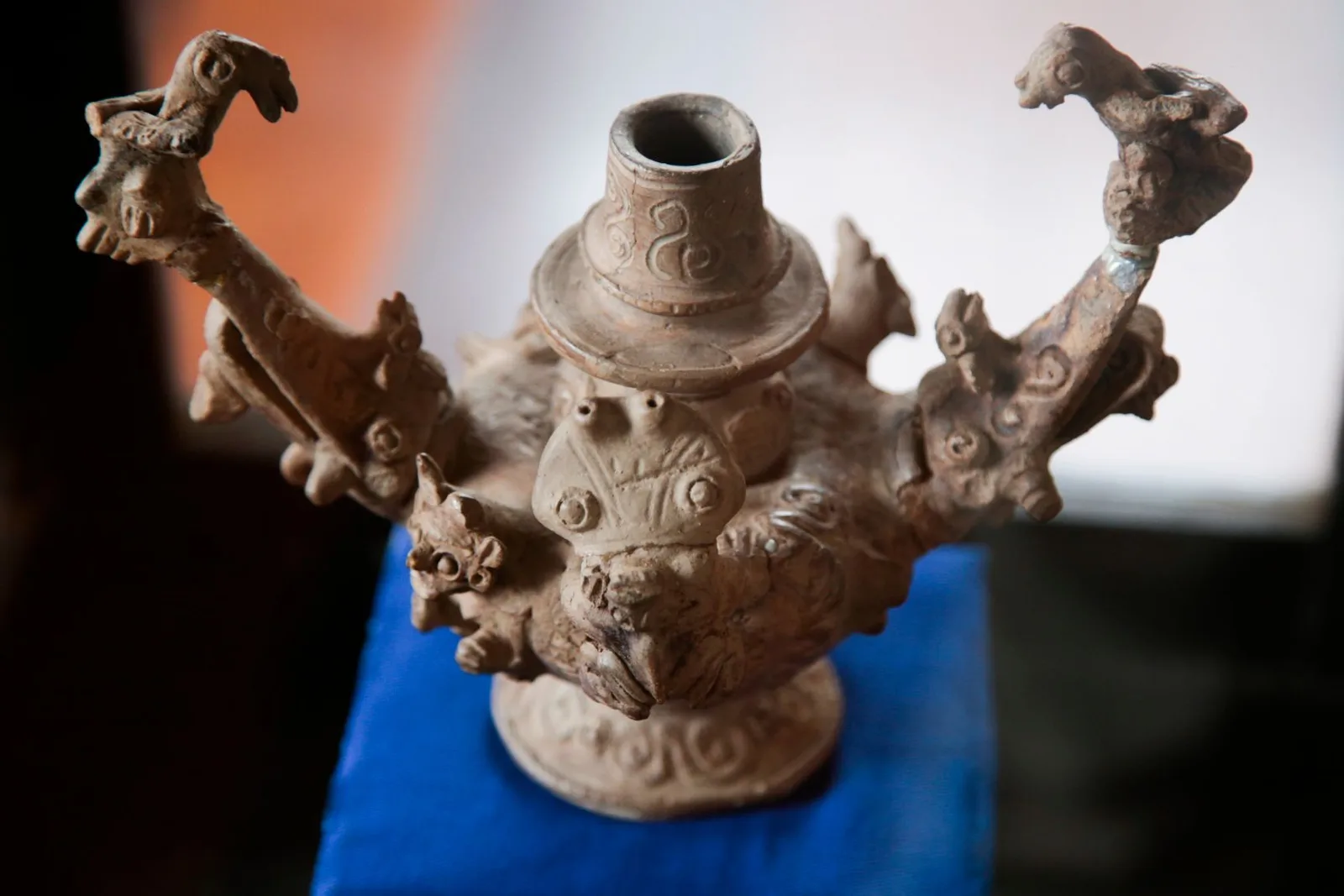 A pale clay vessel with a narrow neck and sculpted animal figures rising from its rim, photographed against a dark background