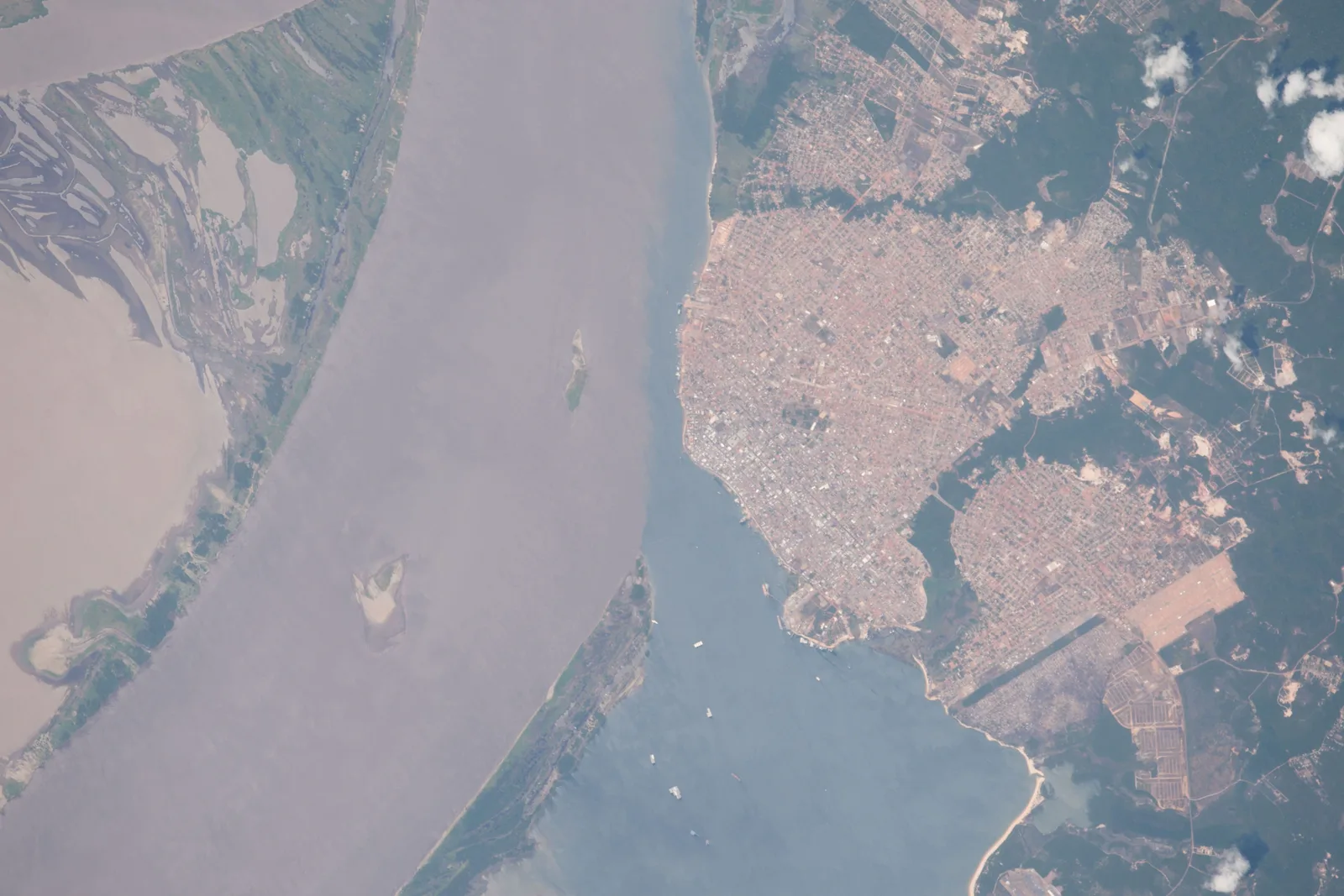 A photograph from the International Space Station: a wide, pale brown river running past the grid of a city on its bank, with the darker water of a second river meeting it