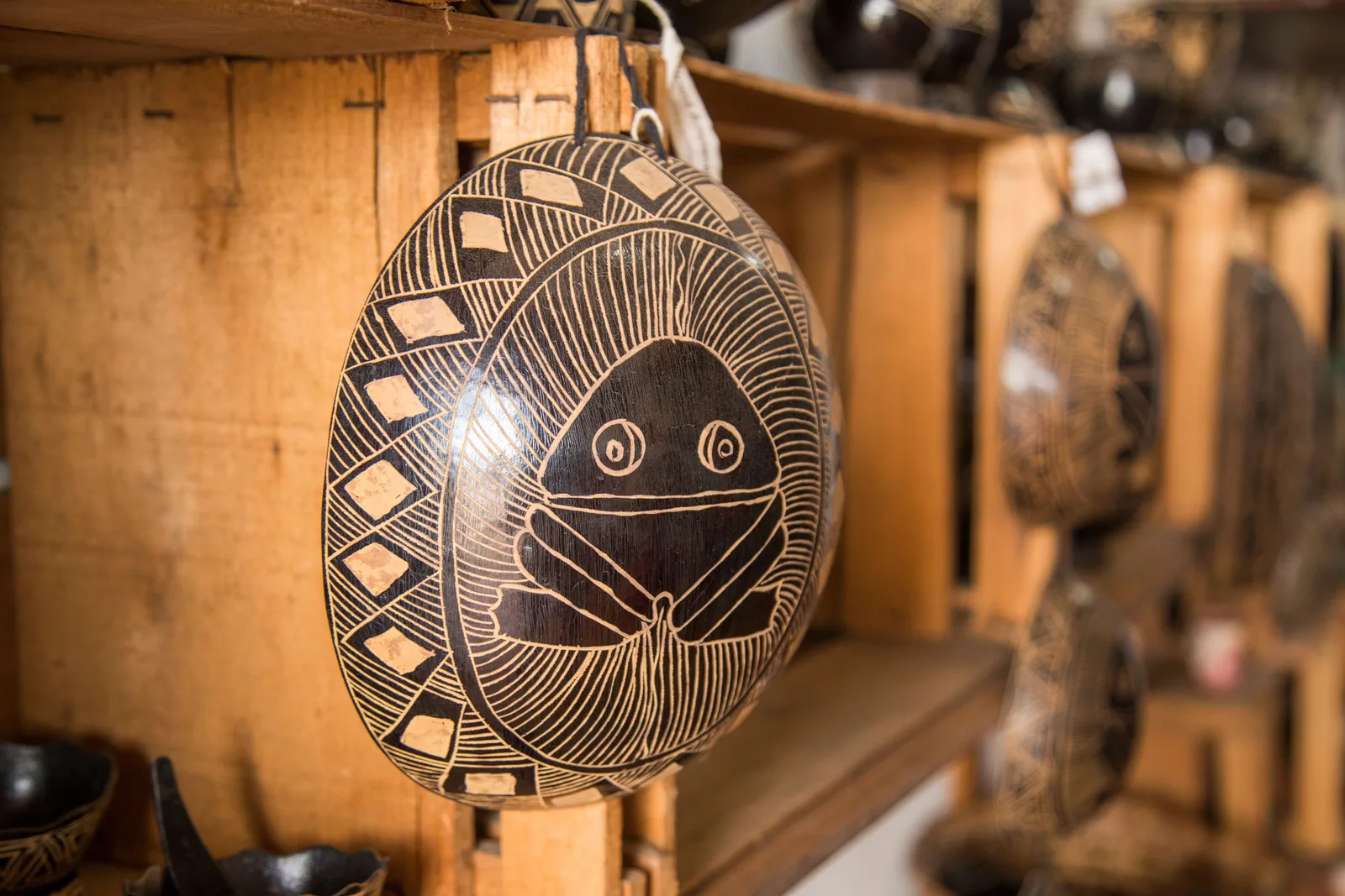 A dark gourd bowl carved with a pale geometric pattern of lines and a stylized face, hanging in a craft shop with more pieces on wooden shelves behind