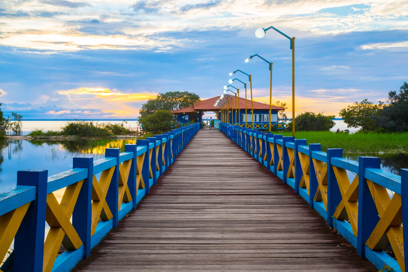 A wooden pier with blue and yellow railings running out over the water to a small roofed shelter, streetlamps along it and a bright evening sky behind