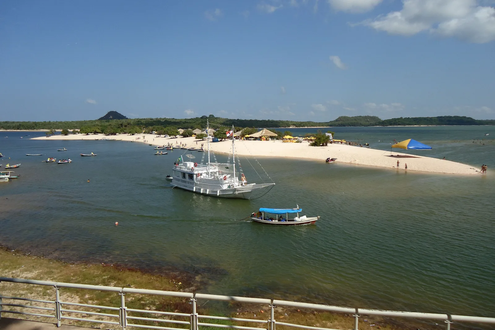 Two white sailboats and a small blue boat moored in green water off a strip of pale sand, a railing in the foreground and a low cone-shaped hill on the horizon