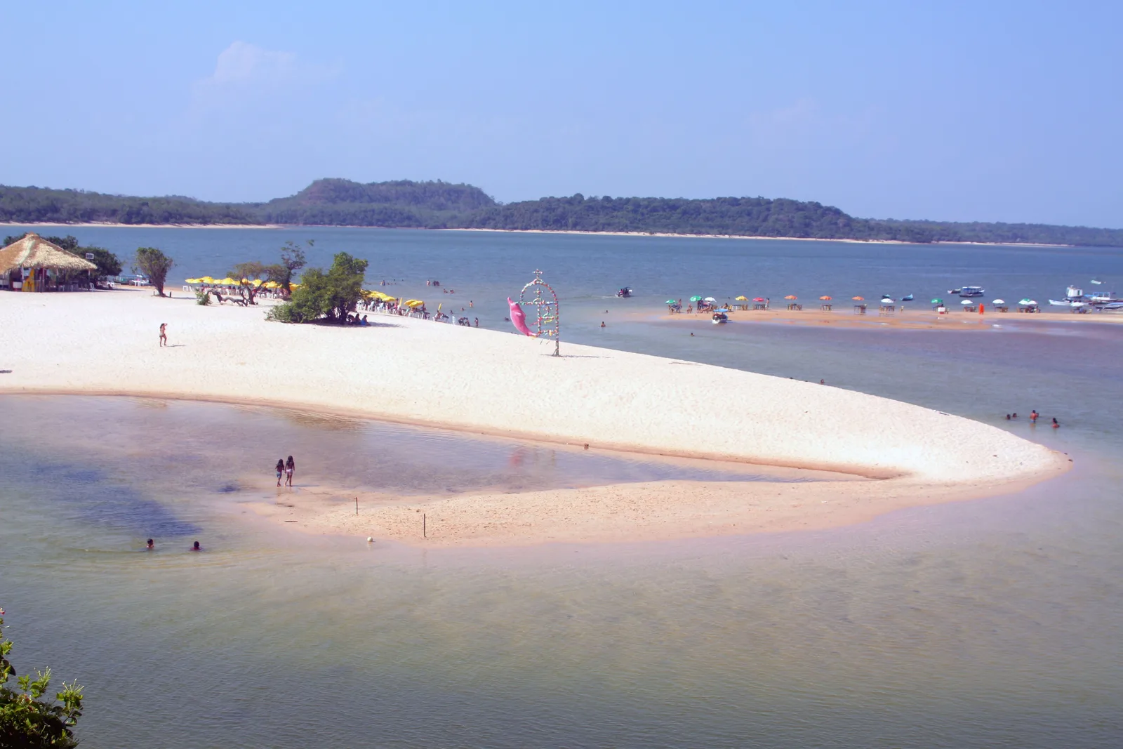 A long, bright white sandbar curving out into shallow river water, people standing far out on it and a line of low flat-topped hills along the far shore