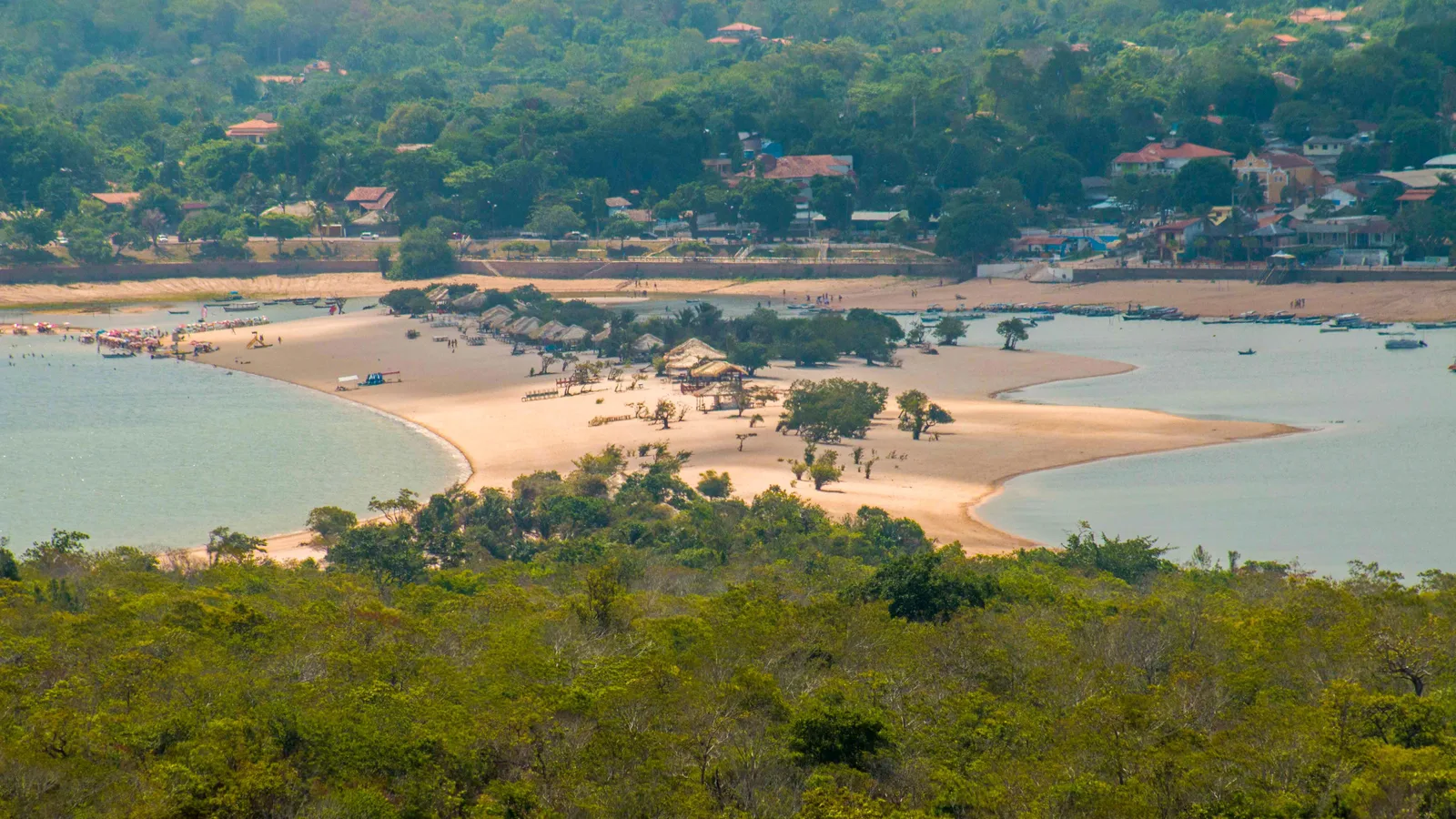 Alter do Chão from the hill across the water: a long curved spit of pale sand with thatched shelters and a few trees on it, pale green river water on both sides, and the village’s red roofs among trees on the far shore