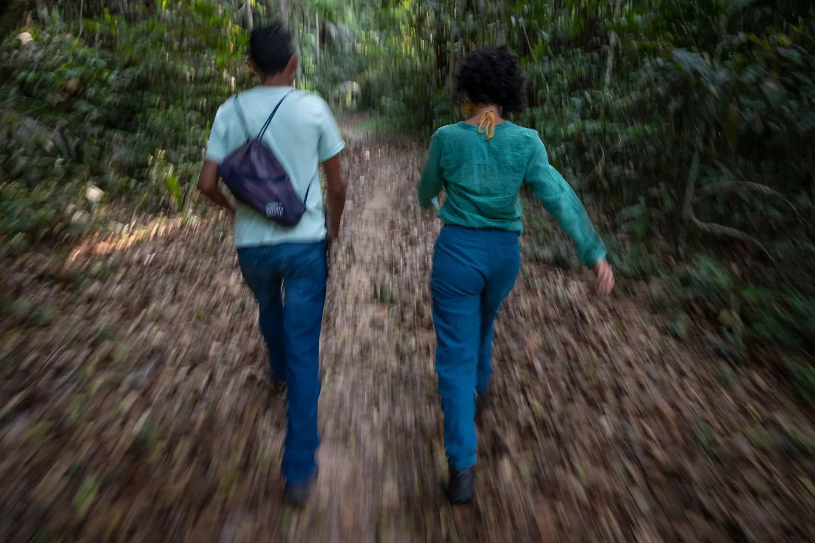 Two people walking away down a narrow trail through dense green forest, one carrying a small backpack