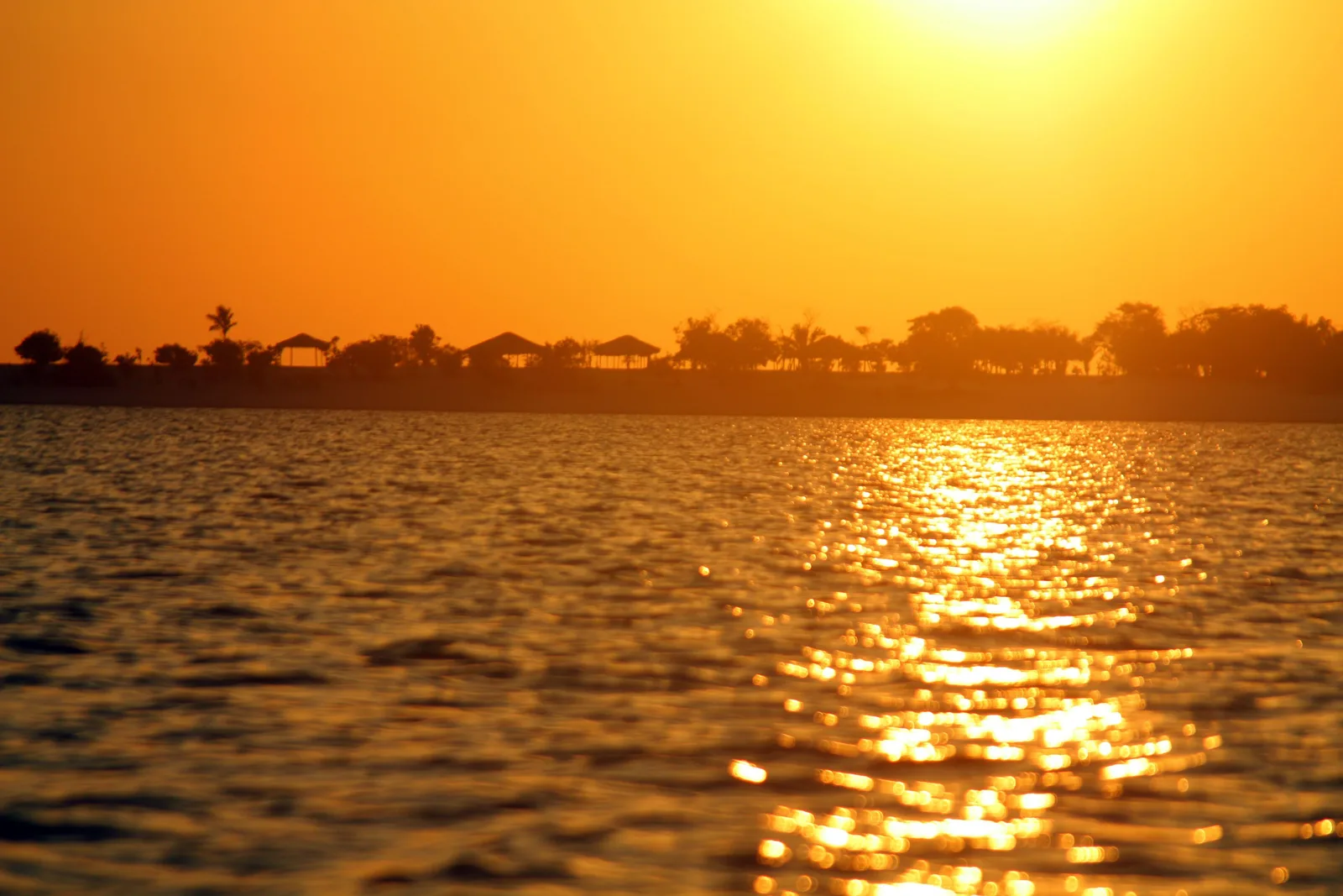 The sun low over a river in a blazing orange sky, a line of trees and thatched roofs silhouetted along the far shore and the water glittering gold