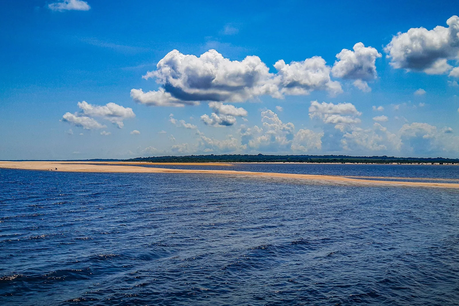 A long, thin line of white sand stretching across deep blue water under a bright sky full of white clouds