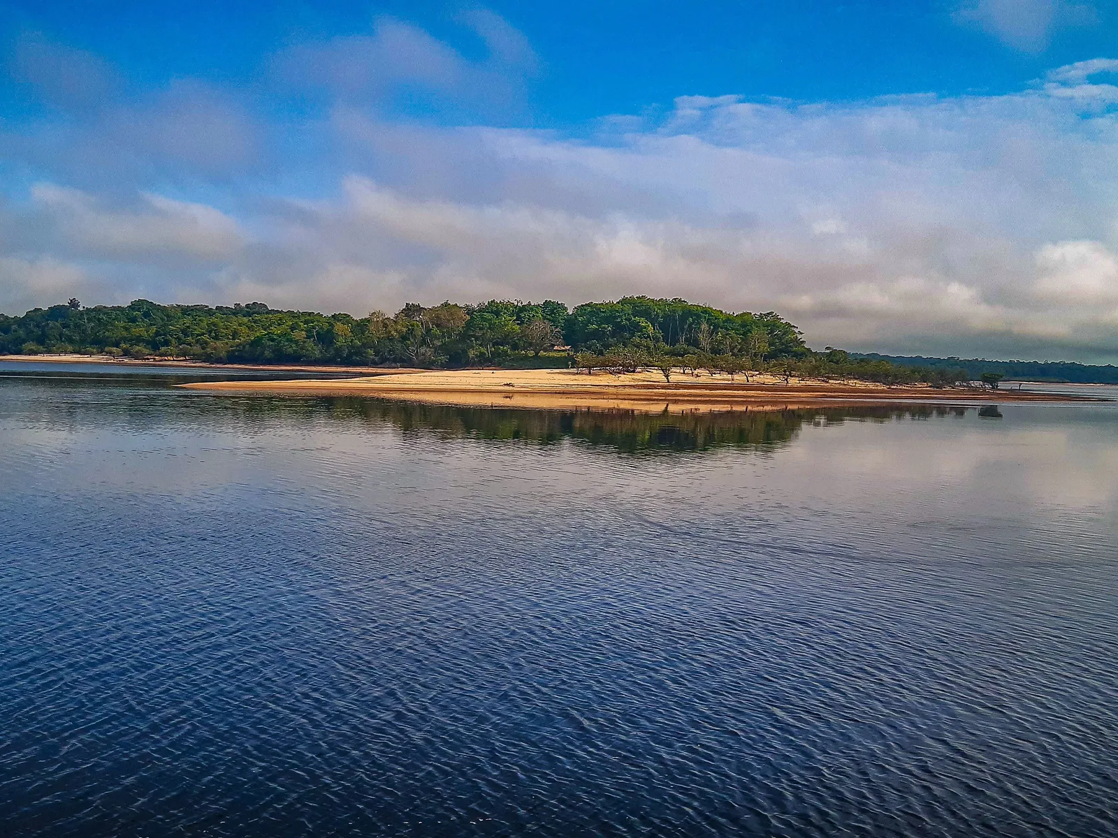 A low wooded island with a white sand beach along its edge, reflected in still blue river water under a morning sky of blue and scattered cloud