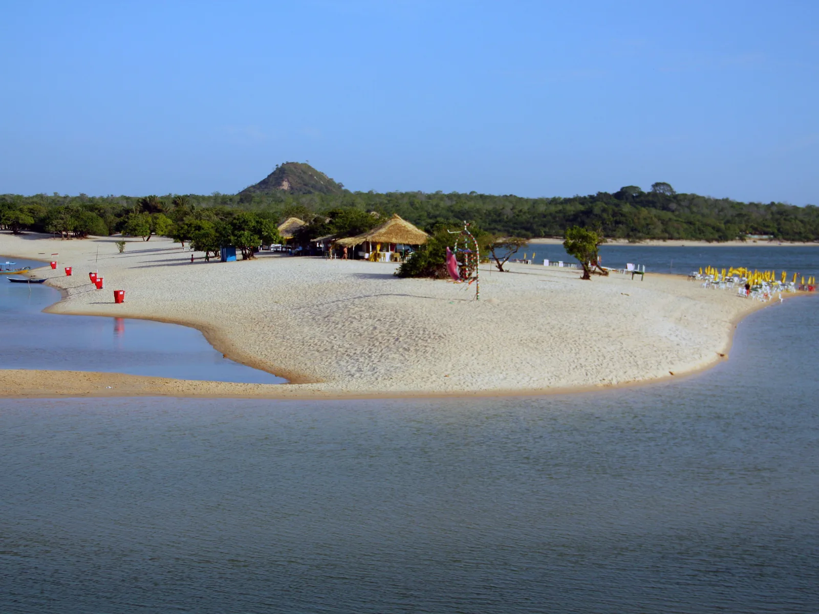 The Ilha do Amor sandbar curving out into calm blue water, thatched shelters and red flags on the sand, and a small cone-shaped hill rising from the forest behind