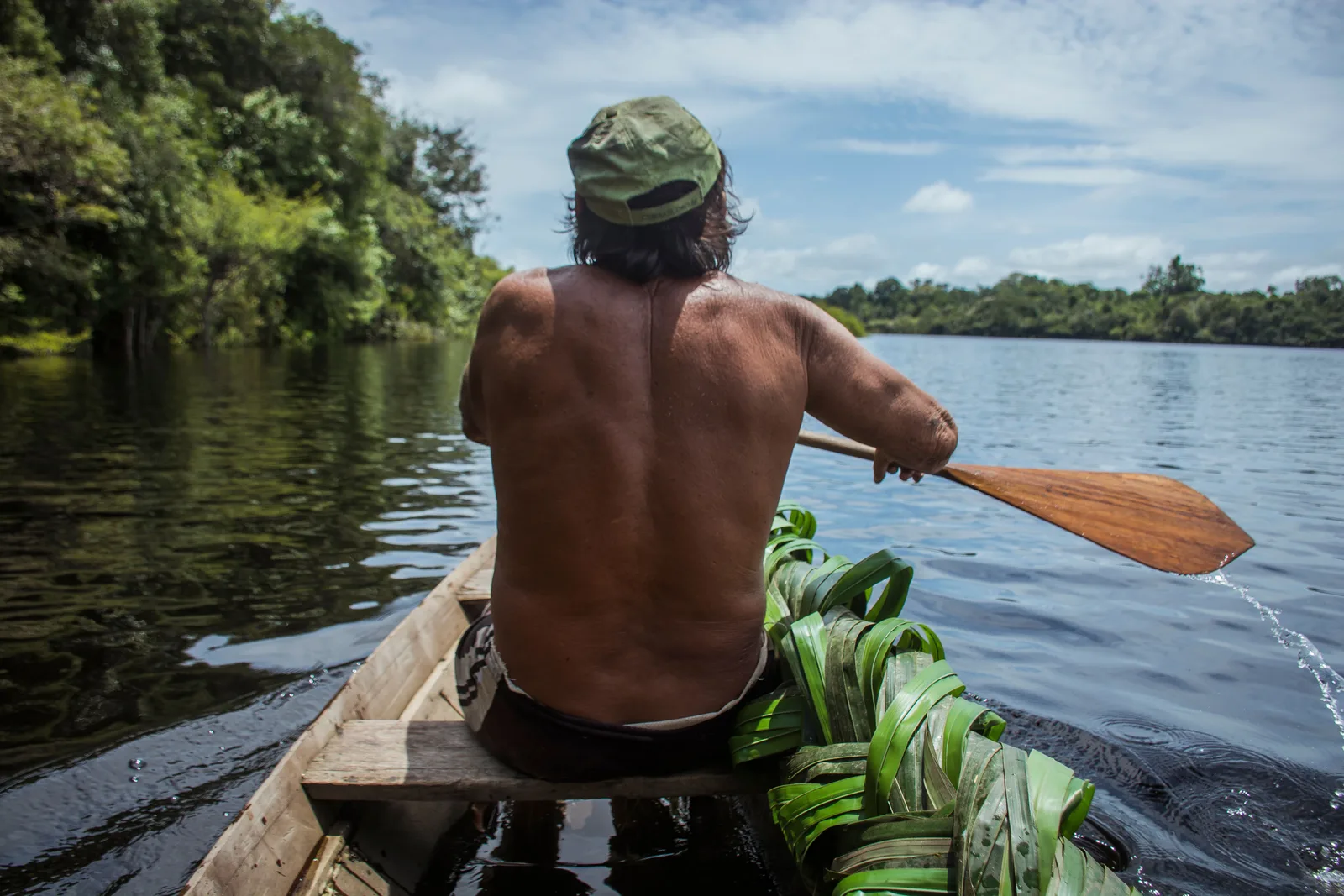 A man in a cap paddling a narrow dugout canoe away from the camera, cut green plants piled in the boat beside him and open river ahead