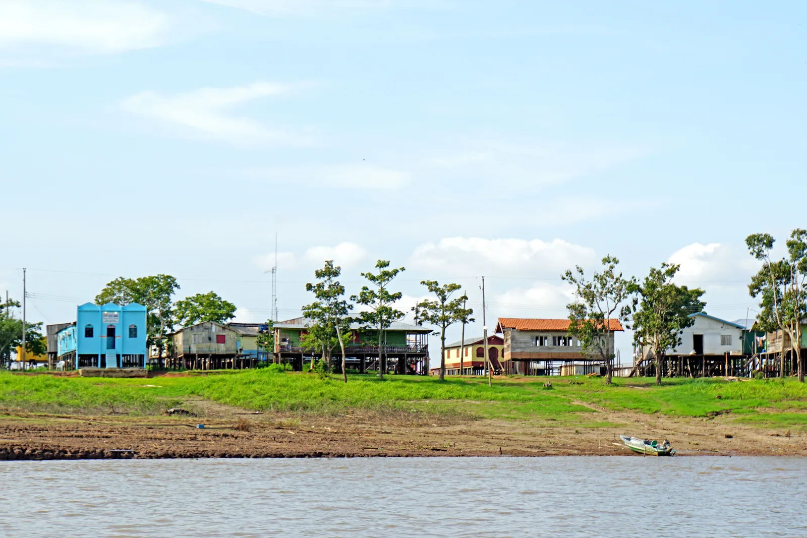 A riverside settlement seen from the water, painted wooden houses spaced along a grass bank above the river