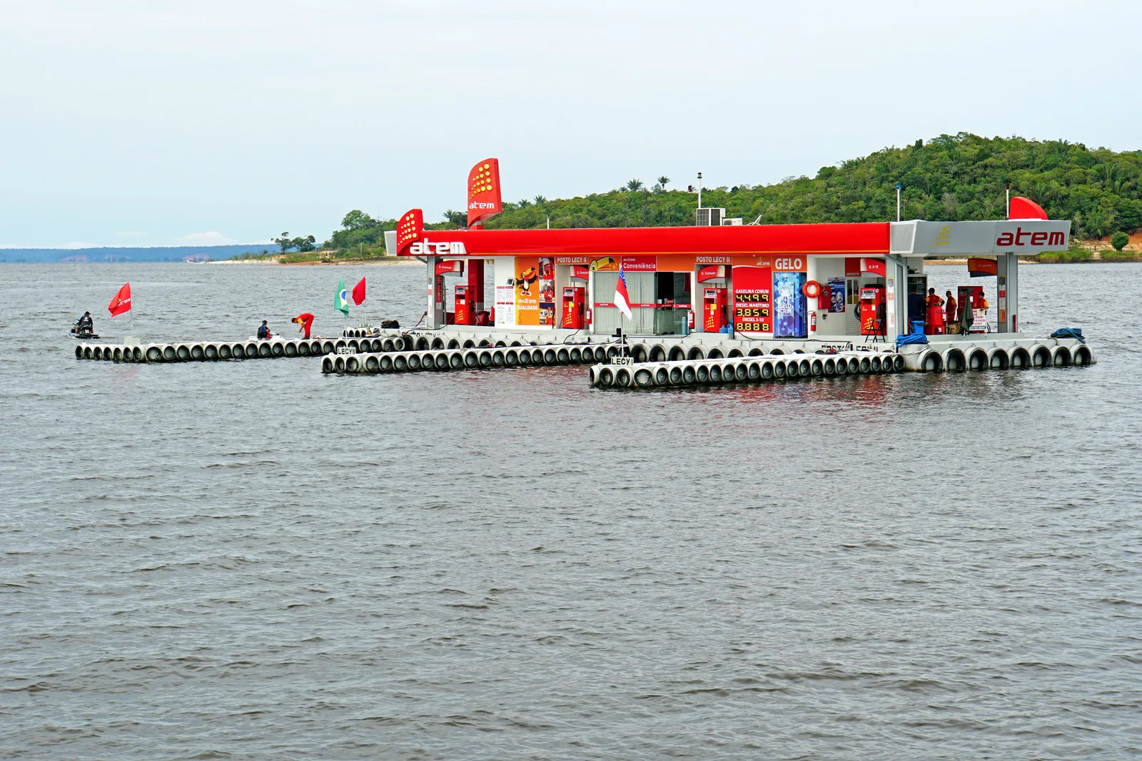 A floating filling station moored out on the river, its red canopies and pumps sitting on a pontoon with forest behind