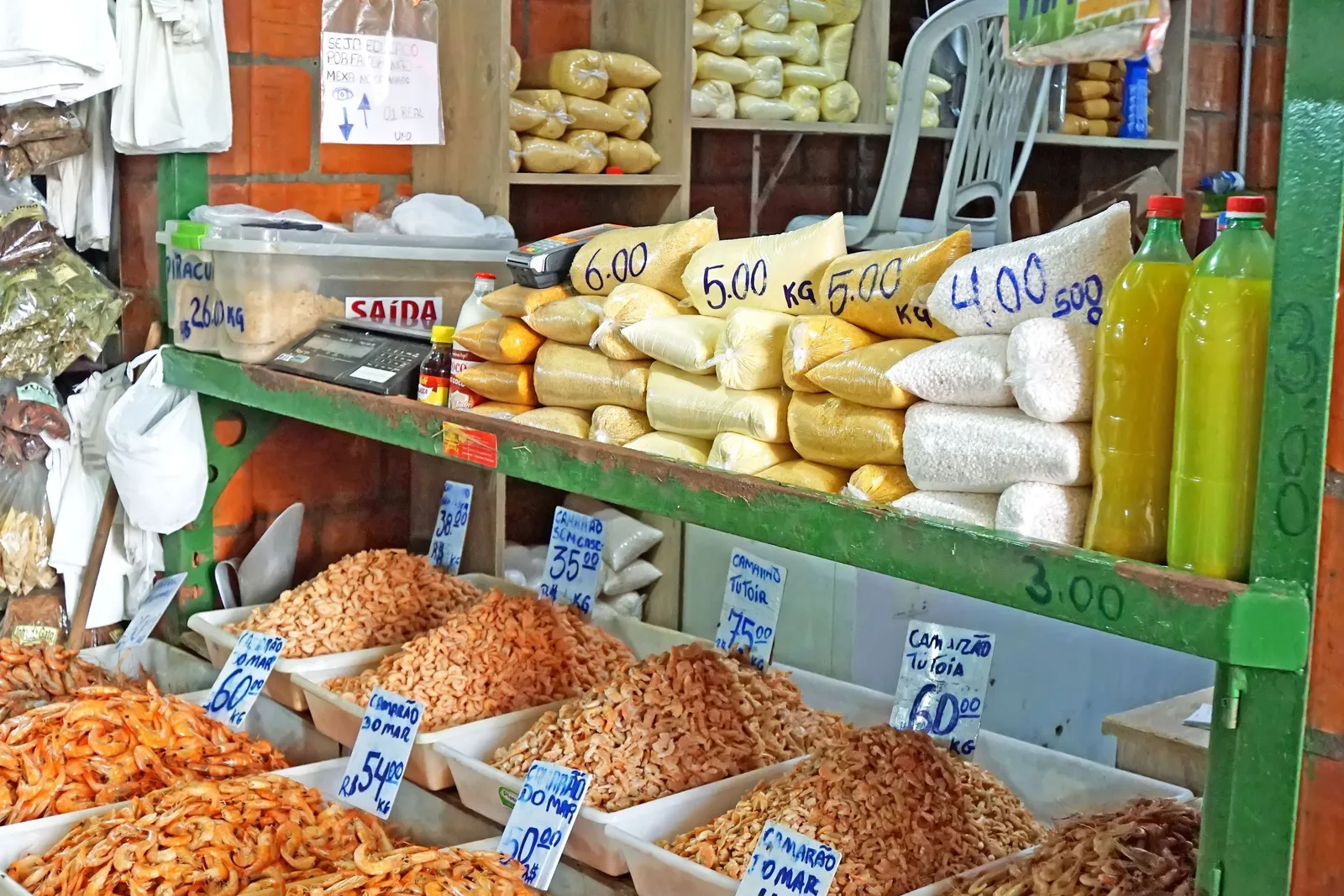 A market stall stacked with bins of dried shrimp and manioc flour and bags of grains, handwritten price cards propped in each one