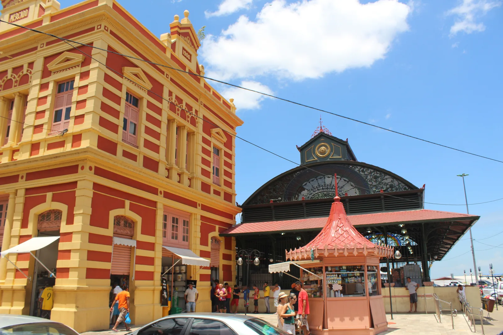 The red and yellow painted facade of the Manaus municipal market, with a dark iron pavilion under an arched roof standing beside it and a small pink kiosk and shoppers in the street in front