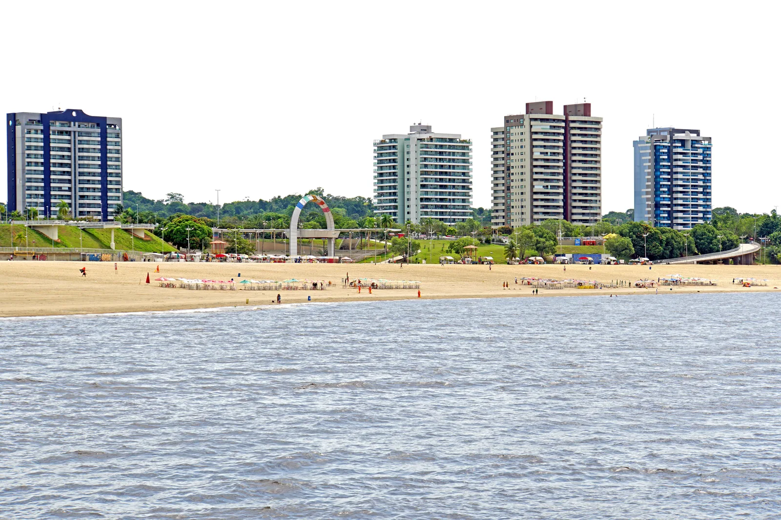 The beach at Ponta Negra seen from the water, people and rows of white parasols along the sand, and four apartment towers — navy, pale green, dark red and blue — standing above the trees behind