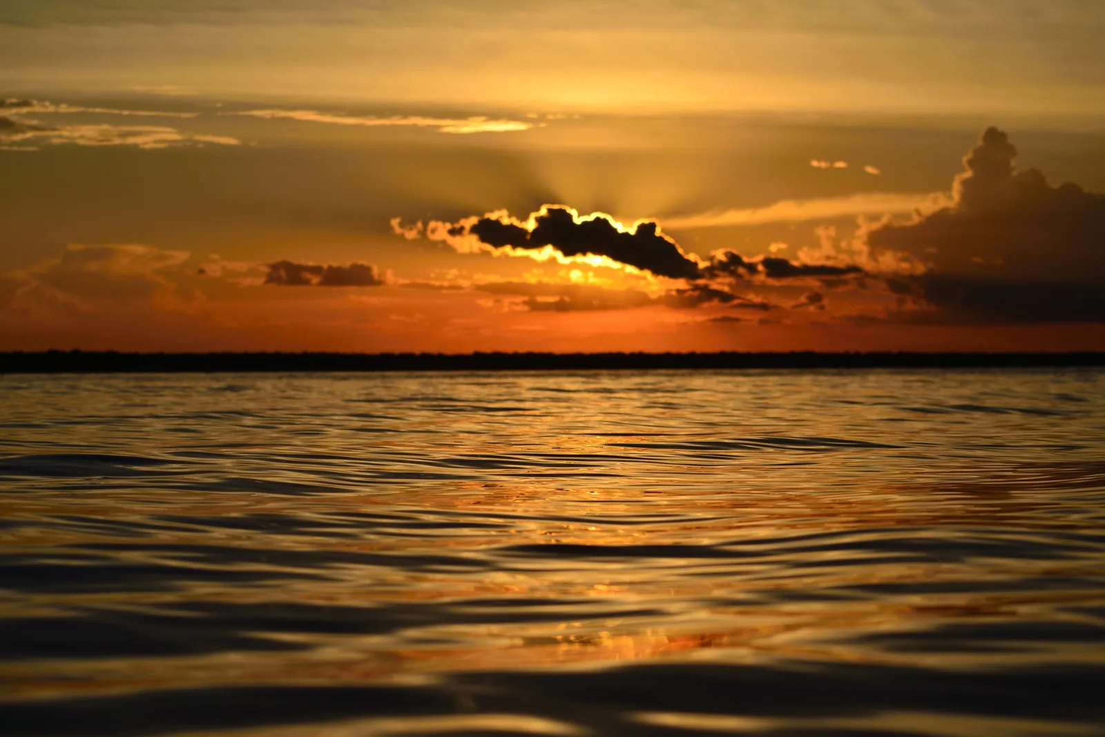 A low orange sun over a wide river with a dark bar of cloud across it and a long track of light running toward the camera