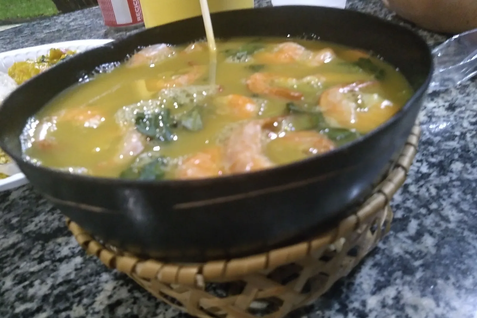 A dark bowl of tacacá in a woven stand, filled with pale yellow broth, pink shrimp and green jambu leaves