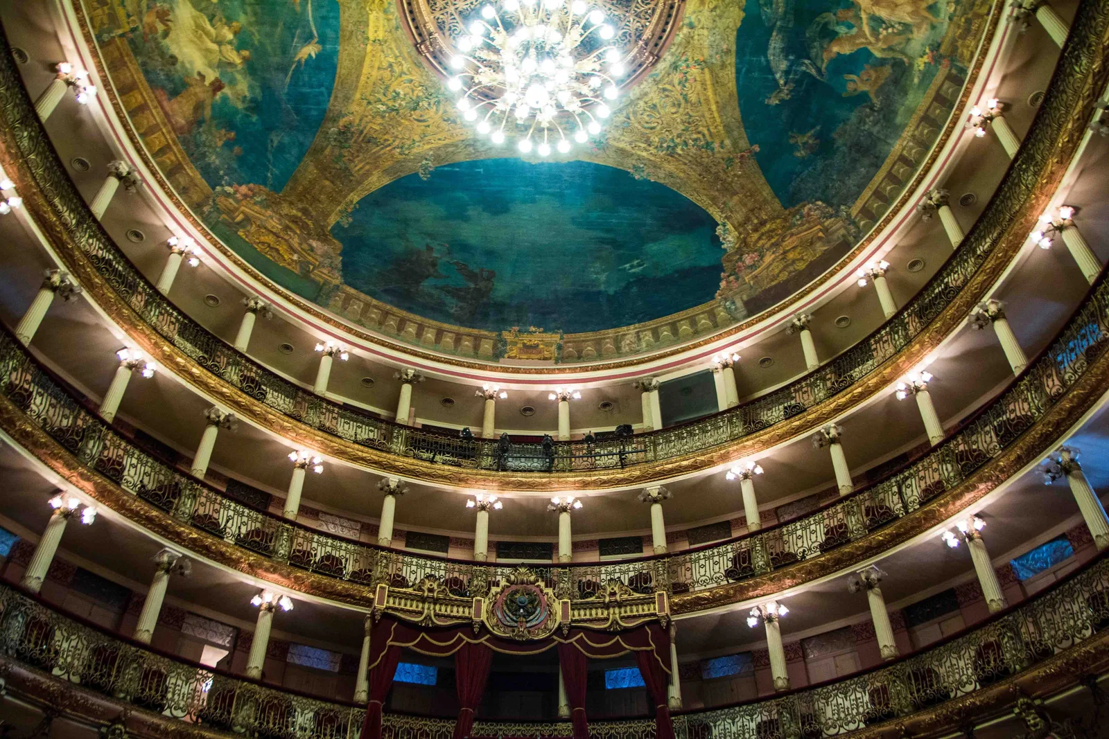 The painted dome of the Teatro Amazonas seen from the floor below, a chandelier hanging at its center above tiers of gilded boxes