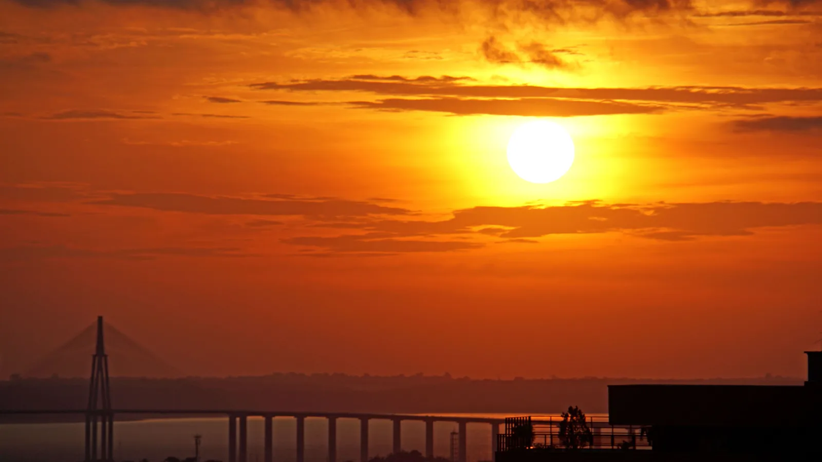 A large orange sun setting over the Rio Negro with the long silhouette of the Manaus bridge crossing the water beneath it