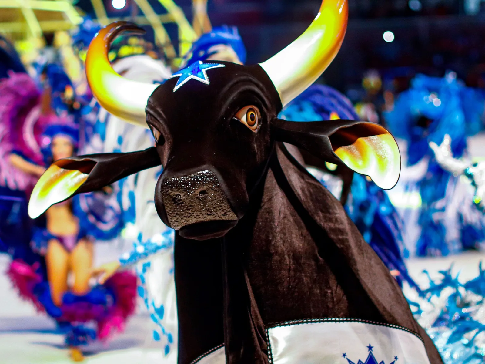 A huge black bull costume with white horns and a decorated head raised above a crowd of performers in blue and white feathered costumes
