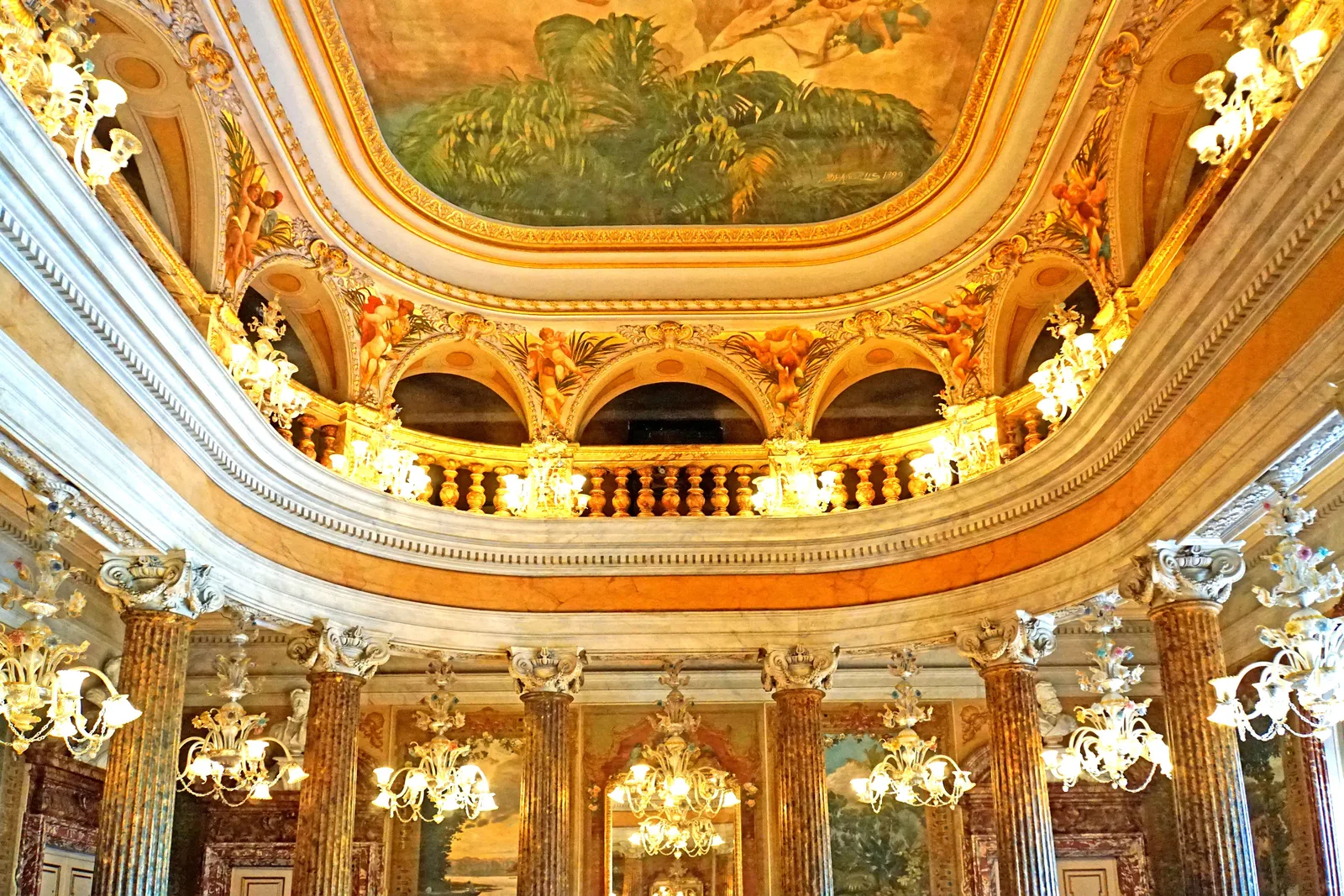 The gilded ceiling of the Teatro Amazonas ballroom, painted panels between columns with chandeliers hanging below