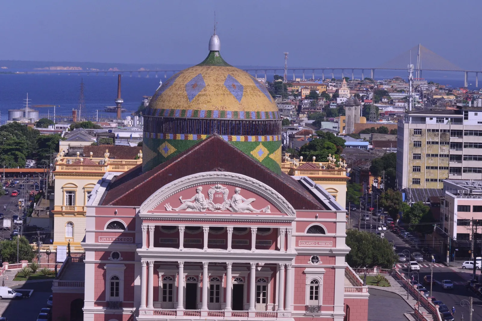 The Teatro Amazonas from above: its pink and cream facade and tiled dome standing among the low buildings of central Manaus, with the river beyond