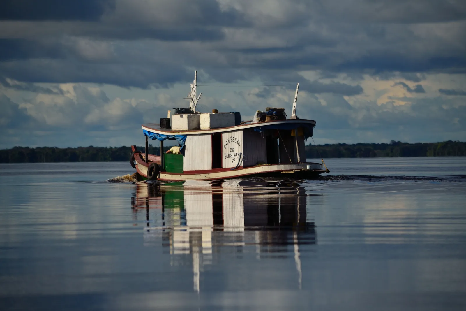 A wooden riverboat sitting on water so still it doubles the hull perfectly, under a flat gray sky