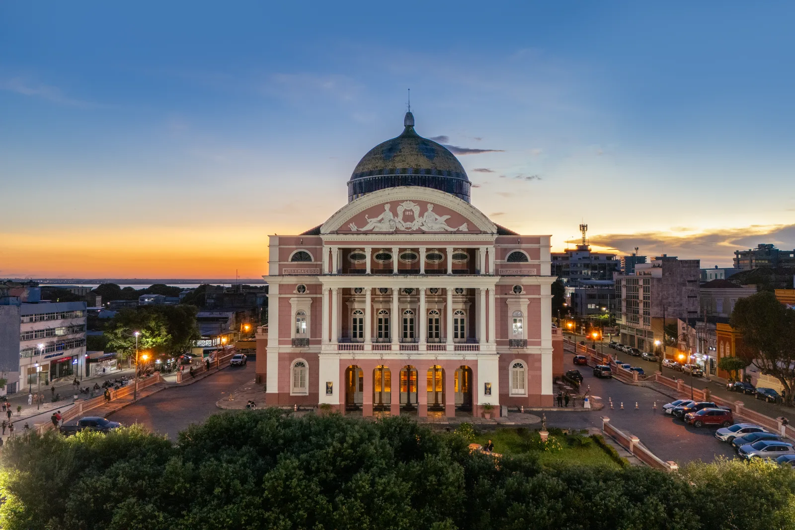 The Teatro Amazonas from above just after sunset: its pink and cream front lit from below, the tiled dome dark against an orange sky, and the streets and buildings of Manaus around it
