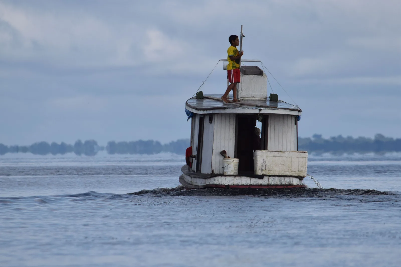 A boy standing on the roof of a small boat crossing a wide gray river, a low green shore in the distance behind him
