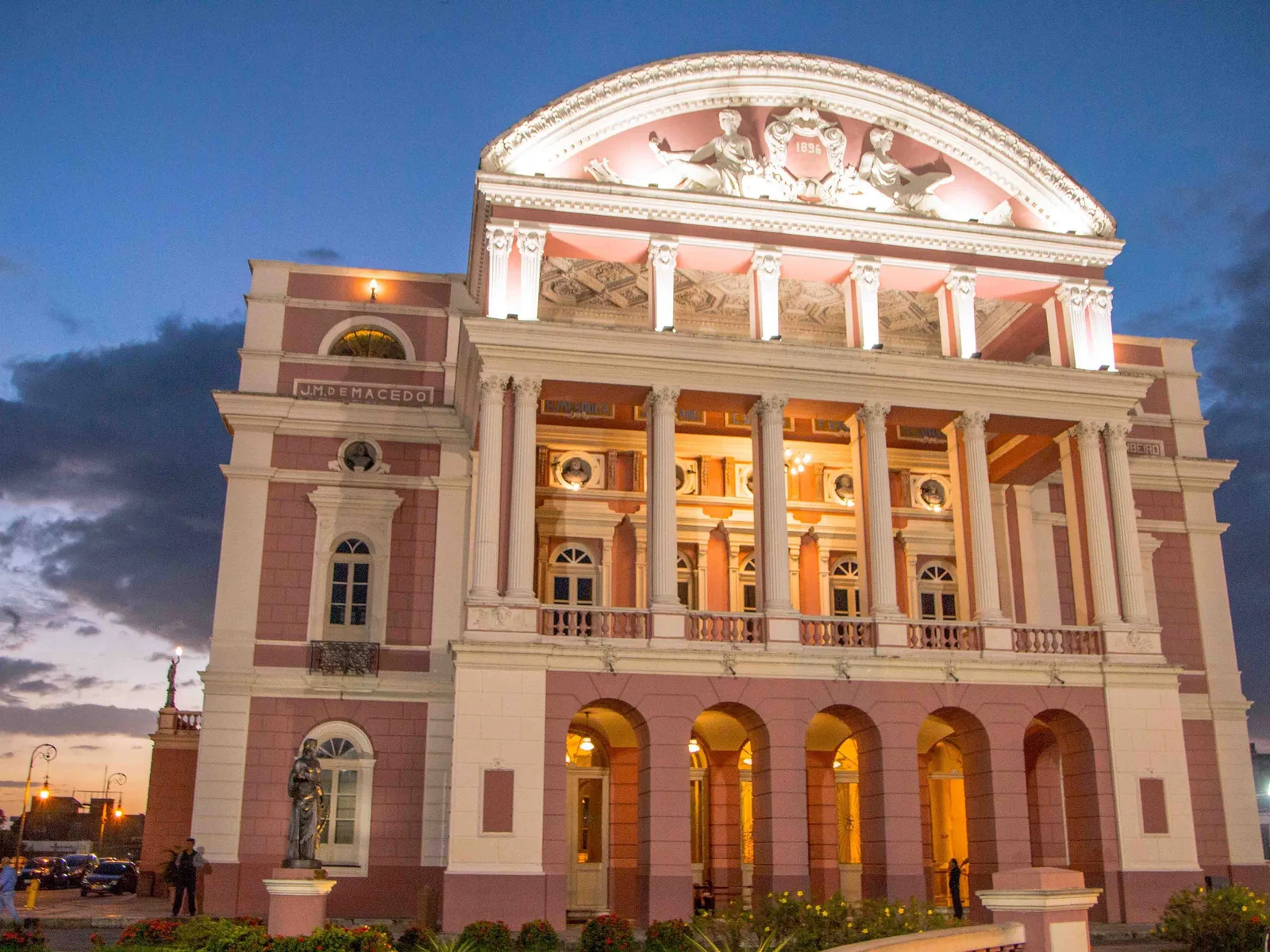 The floodlit front of the Teatro Amazonas at dusk against a deep blue sky, pink and cream stone, a colonnade of tall columns and a sculpted pediment above it