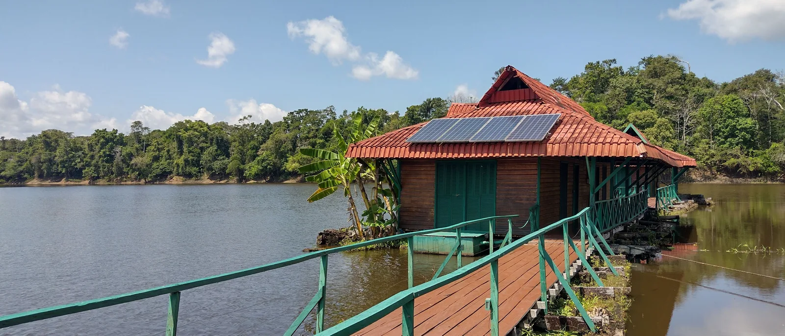 A floating lodge on the river: a green railed walkway leading out to a cabin with a red roof and solar panels, forest closing in behind