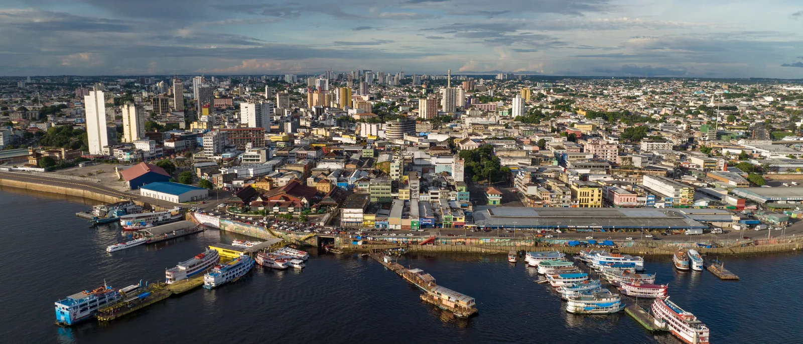 Manaus from across the river: riverboats moored in ranks along the shore below a dense city of low buildings and scattered tower blocks